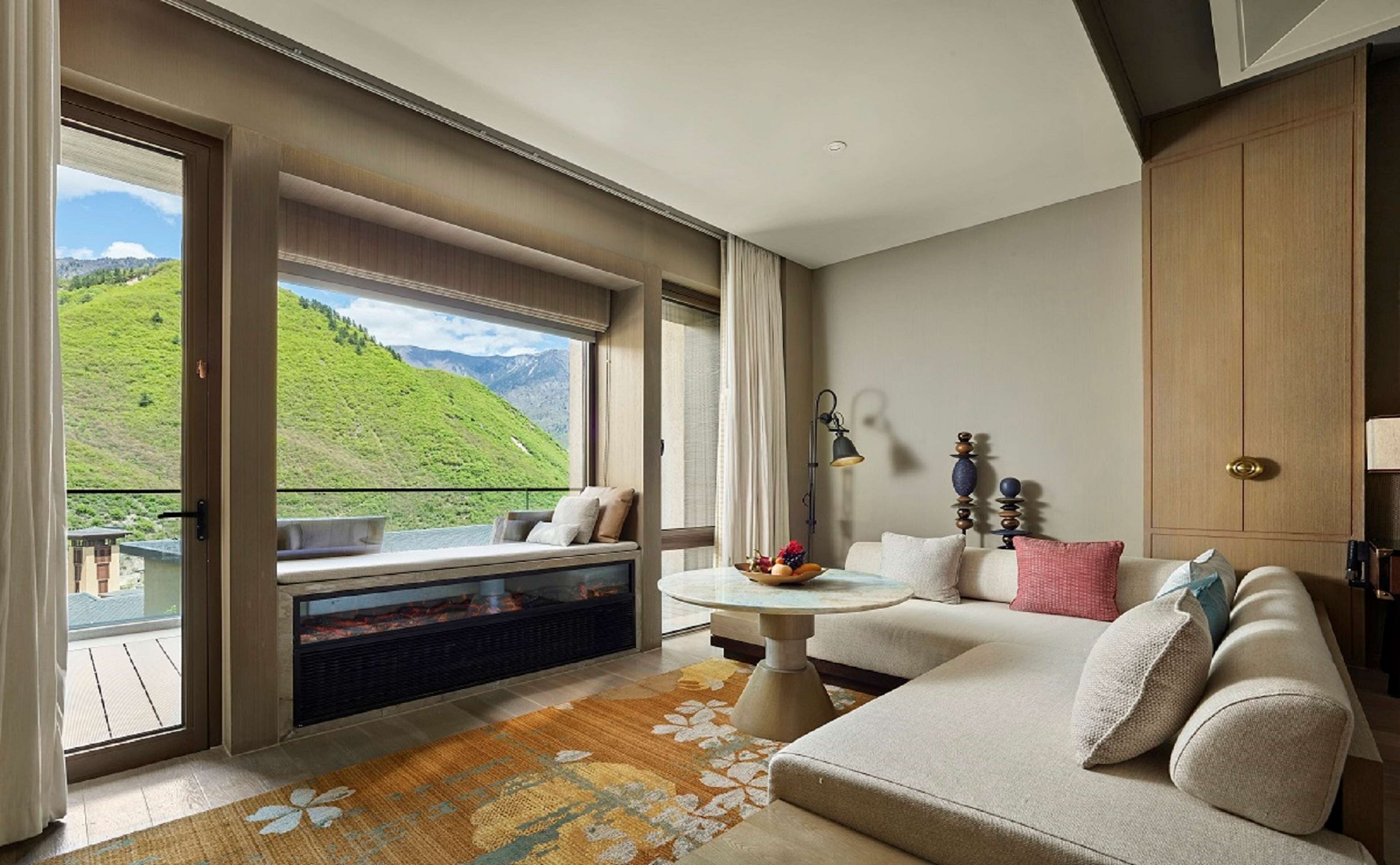 A modern hotel room with a large window offering a mountain view, a cozy seating area with a sofa and a small table, and a wooden wardrobe.