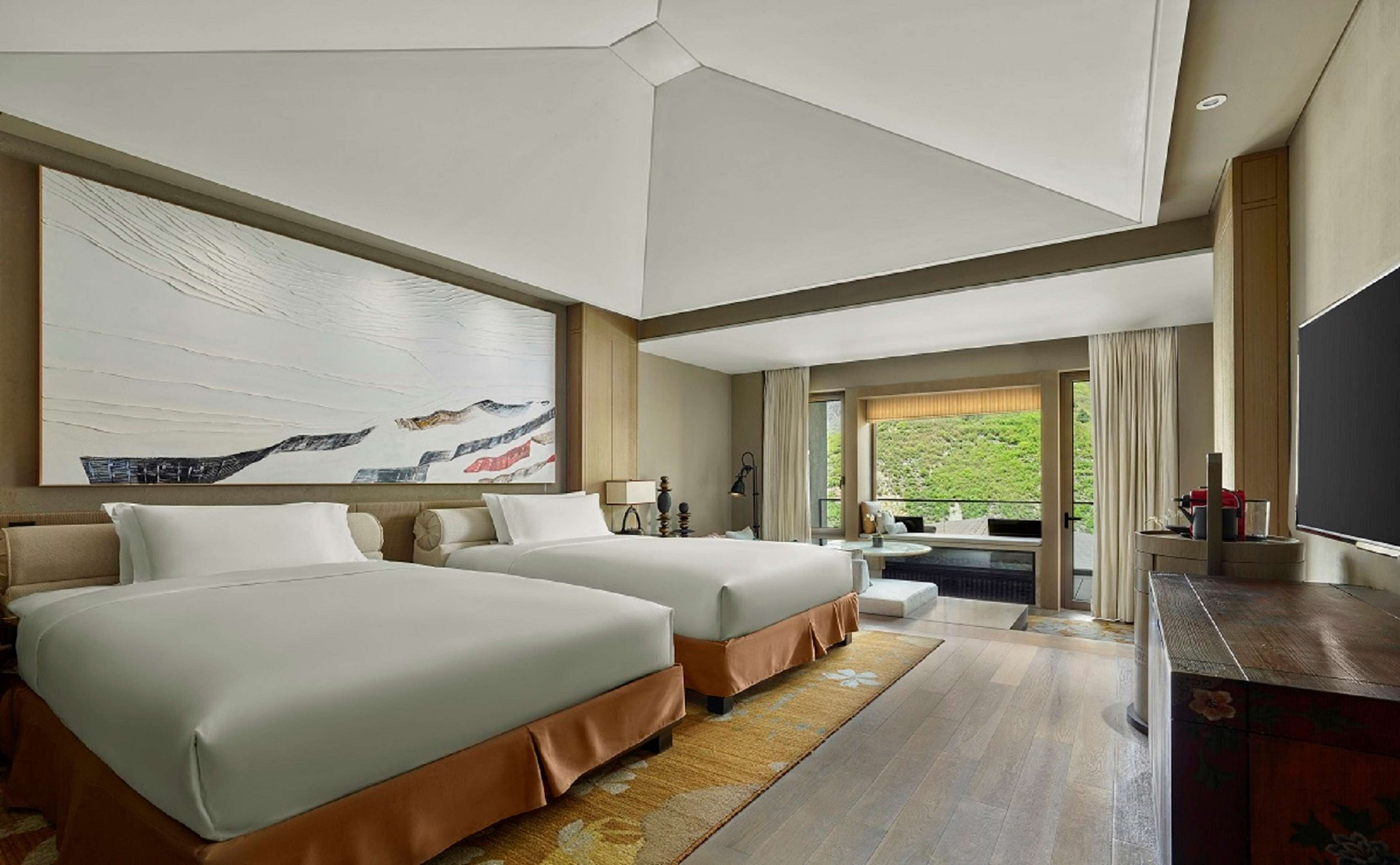 A hotel room with two beds, a large abstract painting, a television, and a view of greenery through the window.