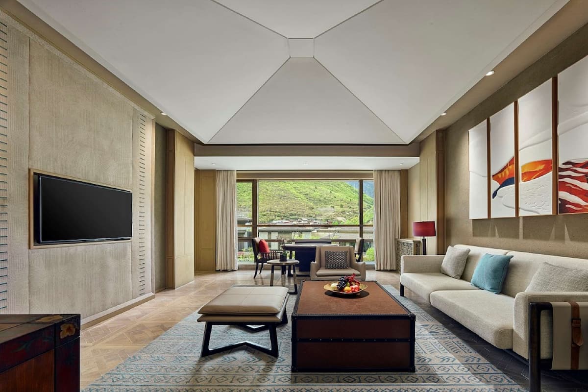 A spacious living room with a large TV, a sofa, a coffee table, and a view of a green landscape through large windows.