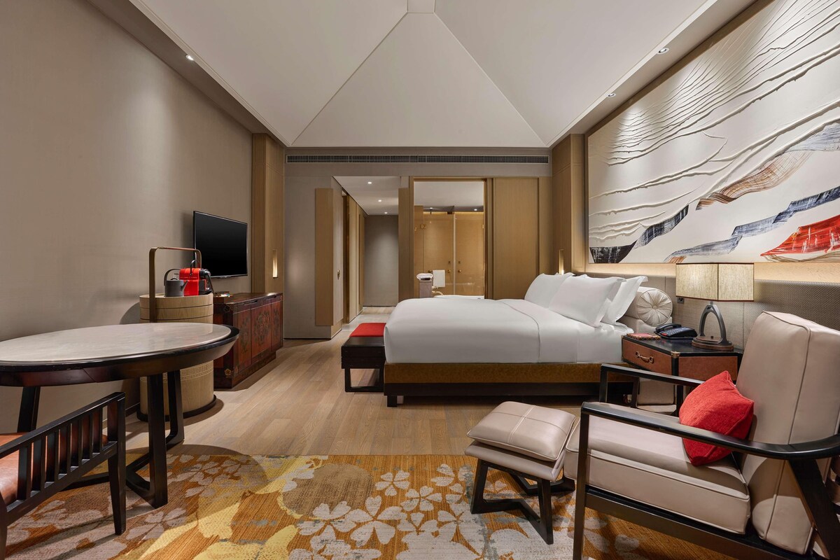 A modern hotel room with a large bed, a desk with a television, a seating area with a chair and ottoman, and a decorative wall mural.