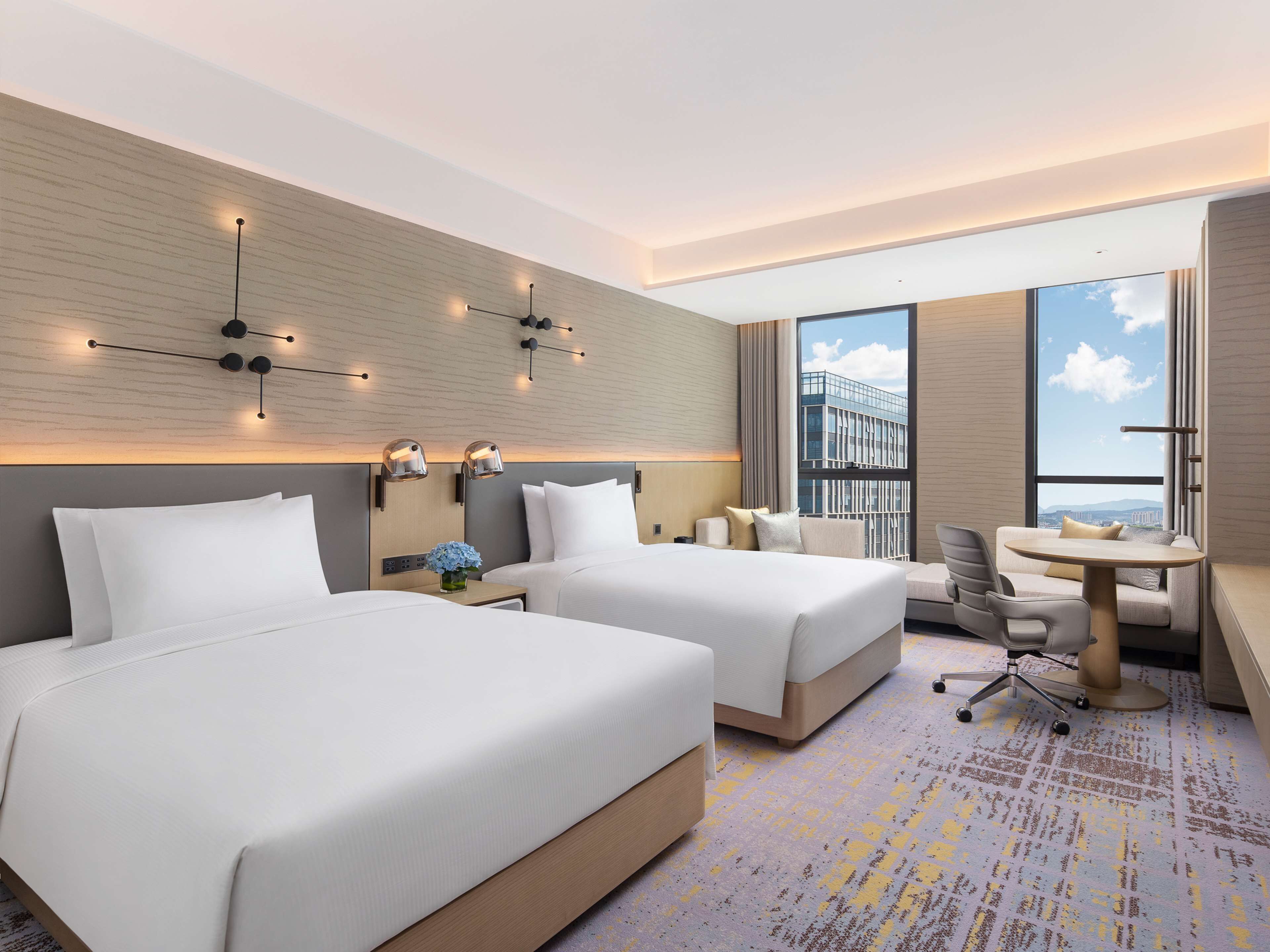 A hotel room with two beds, a desk, a chair, and a large window offering a city view.