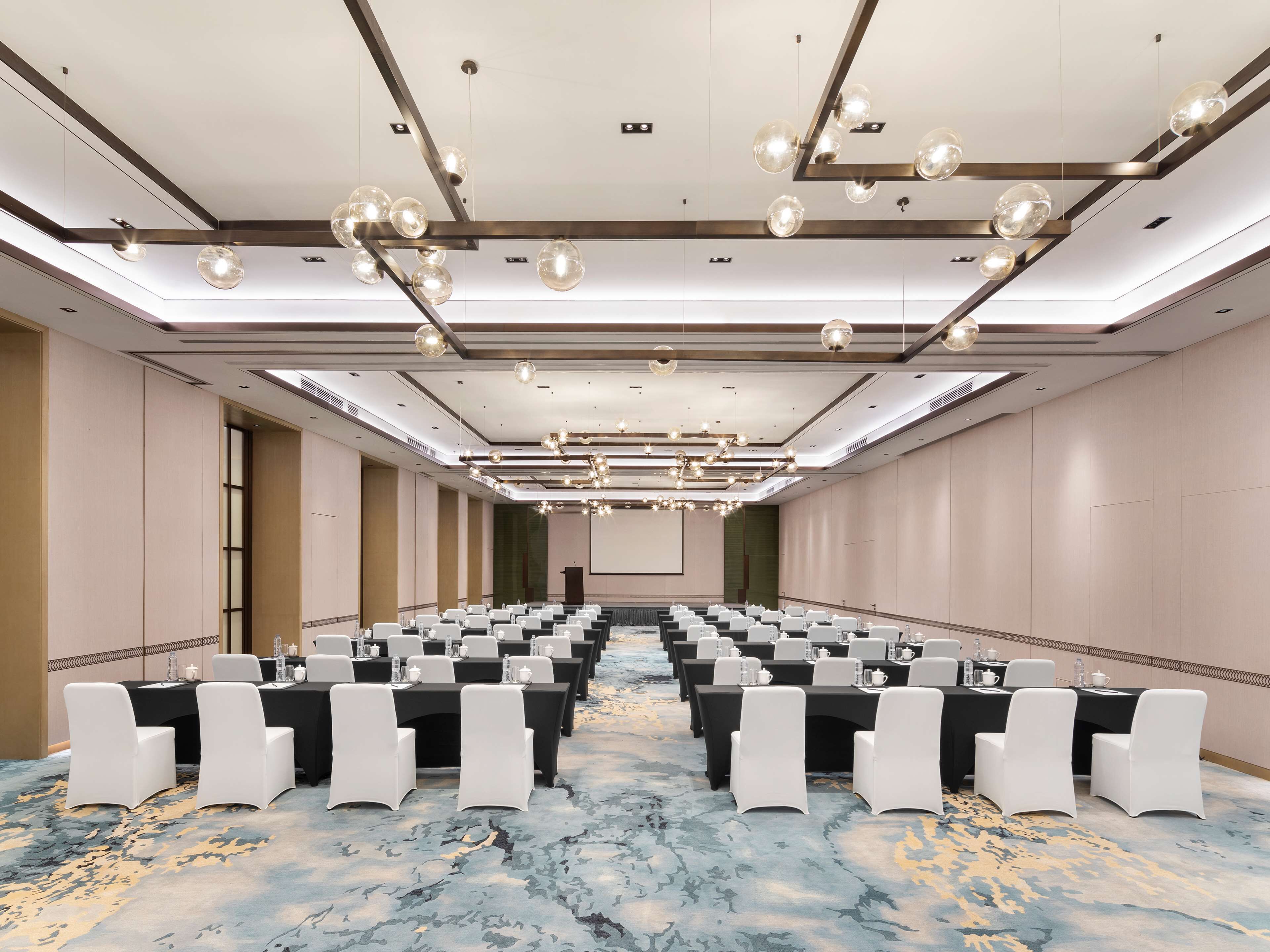 Un salón de conferencias con filas de mesas y sillas, una alfombra estampada y un diseño de techo moderno.