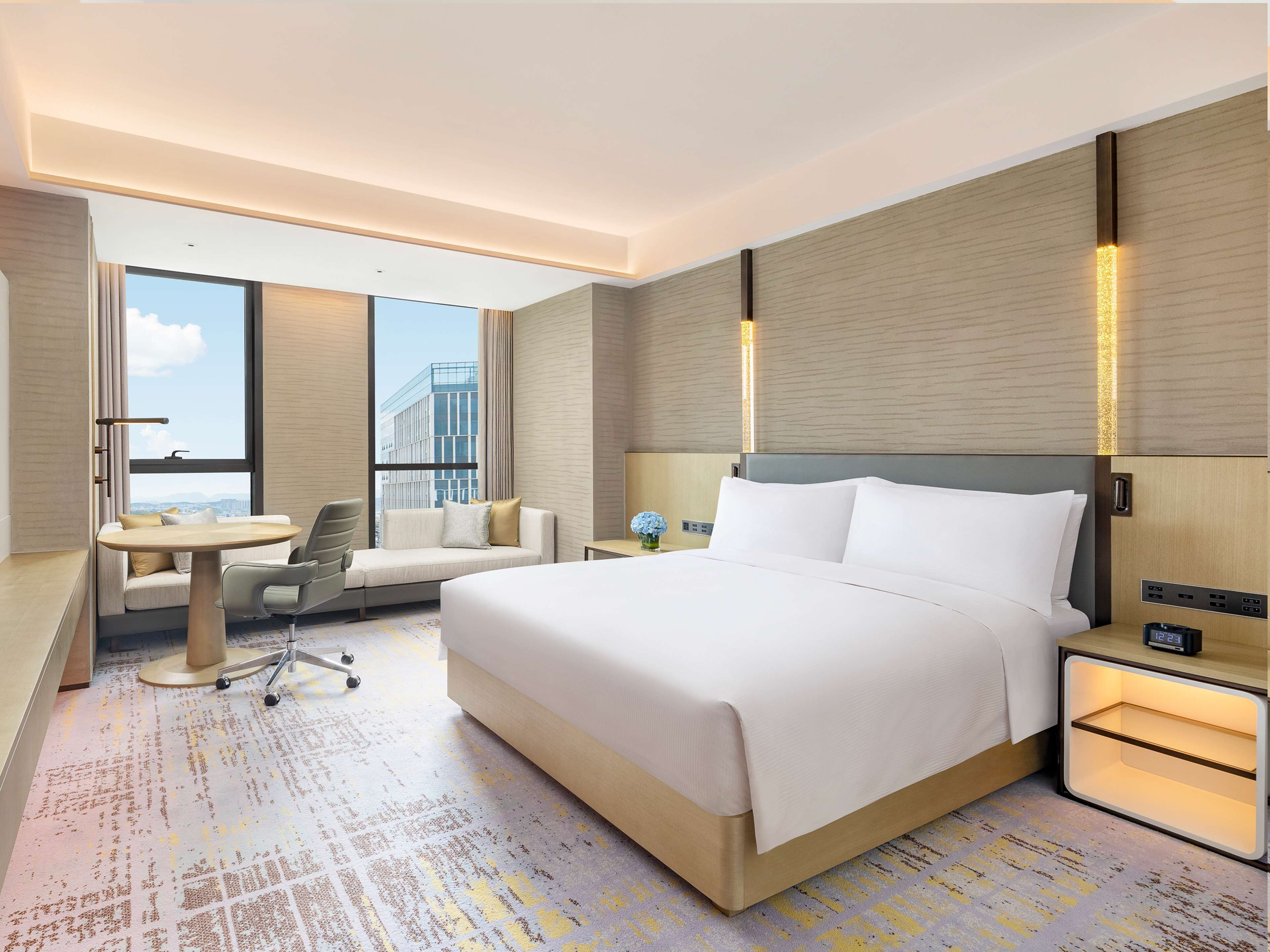 A modern hotel room with a large bed, a sofa, a desk, and a view of the city.