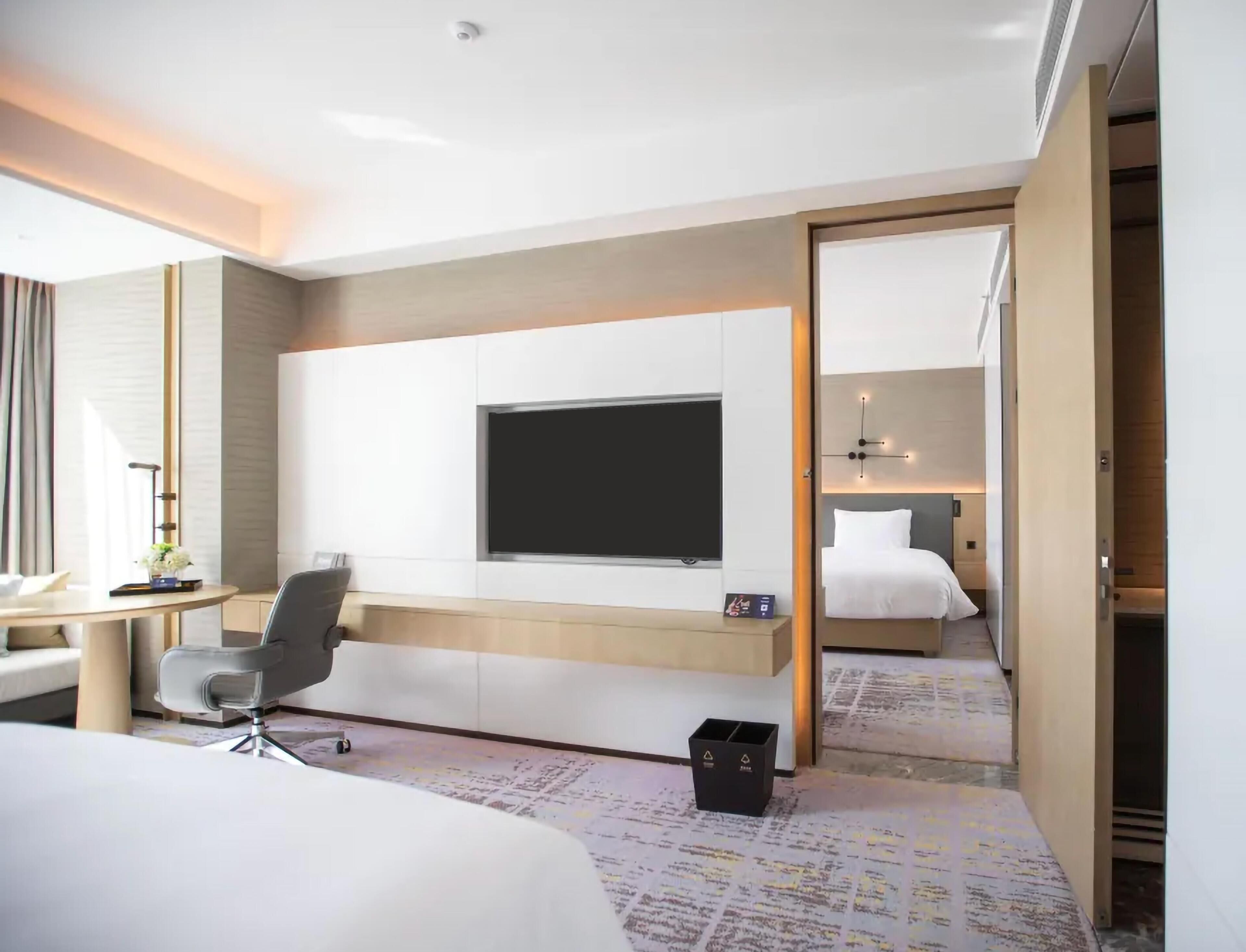 A hotel room with a bed, a desk, a chair, a TV mounted on the wall, and a wardrobe.