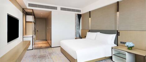 A modern hotel room with a large bed, a bedside table with a vase of flowers, a flat-screen TV, and a walk-in closet.