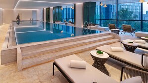 Una piscina interior moderna con paredes de vidrio transparente, áreas de descanso con sofás y sillones, y vistas a la ciudad a través de amplios ventanales.