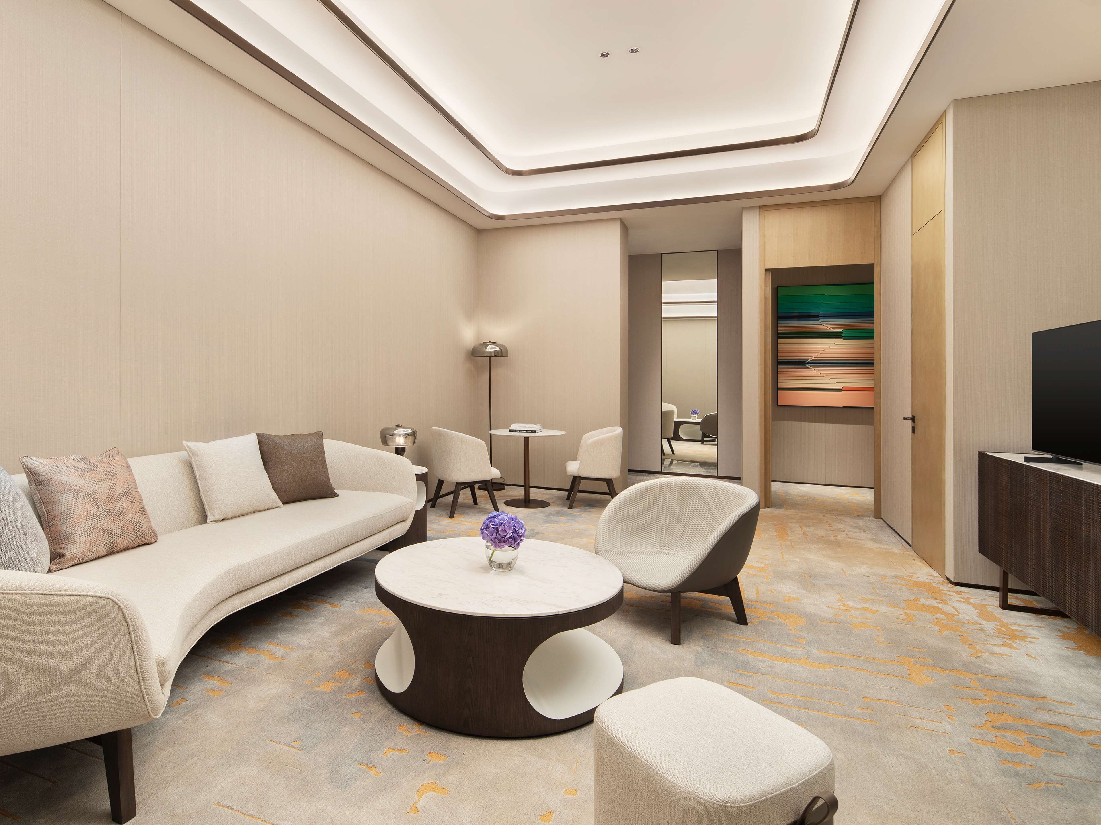 A modern hotel room with a curved sofa, a round coffee table, a chair, a small ottoman, and a TV on a cabinet.