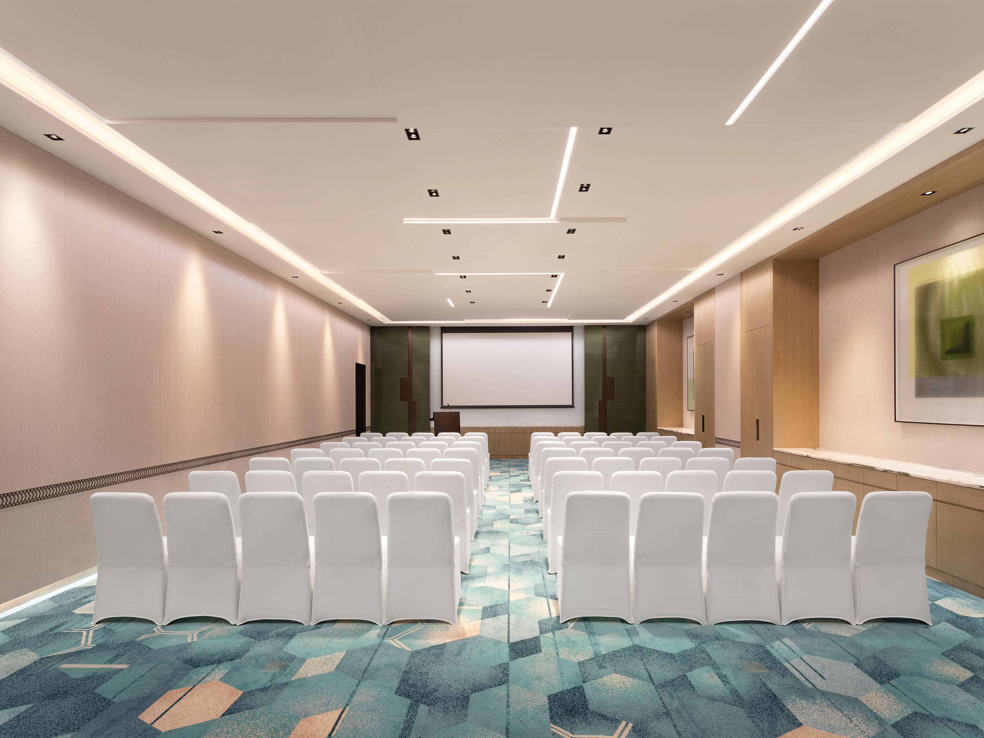 A conference room with rows of white chairs, a projector screen, and a carpeted floor.