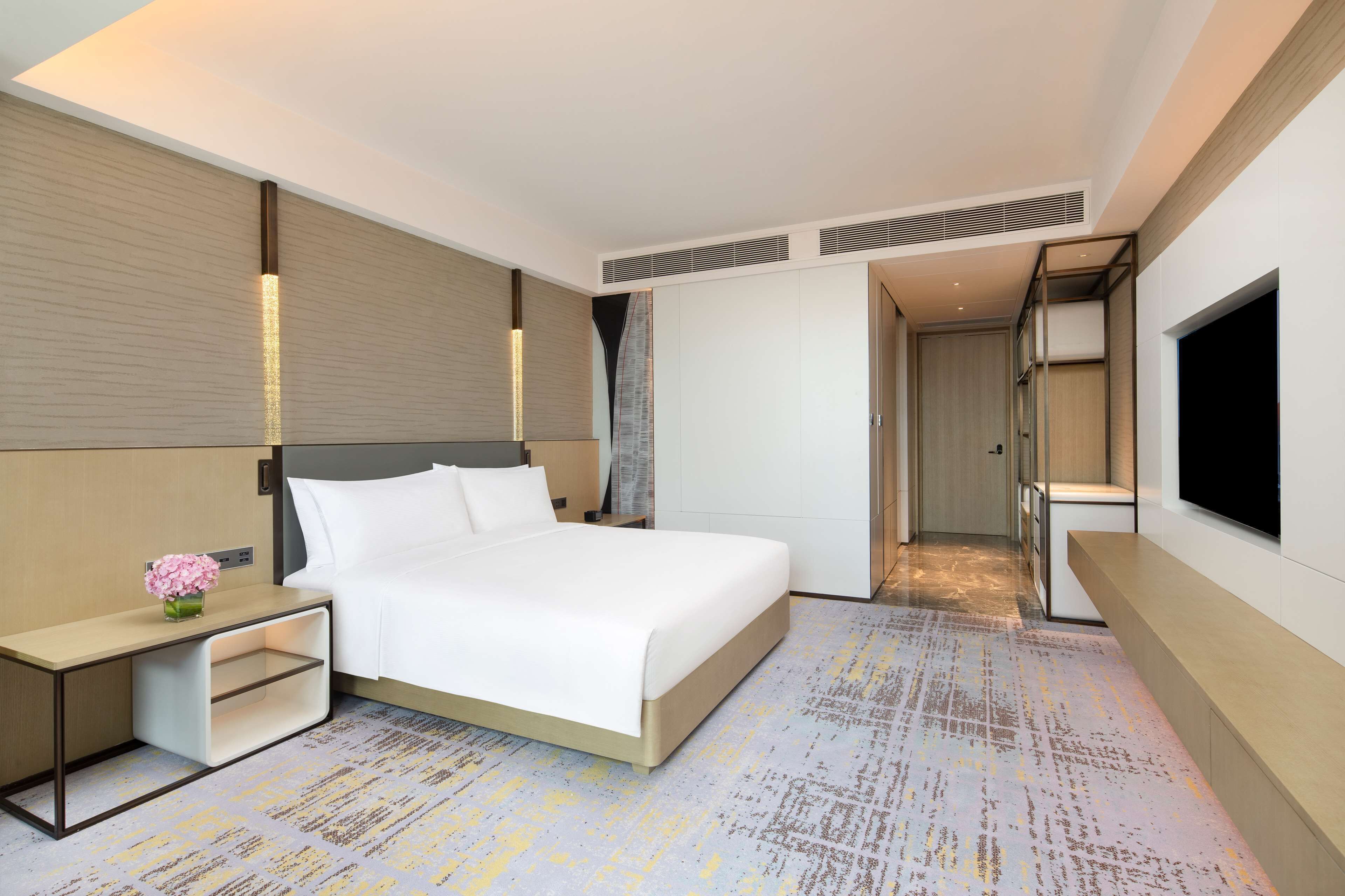 A modern hotel room with a large bed, bedside tables, a flat-screen TV, and a wardrobe.