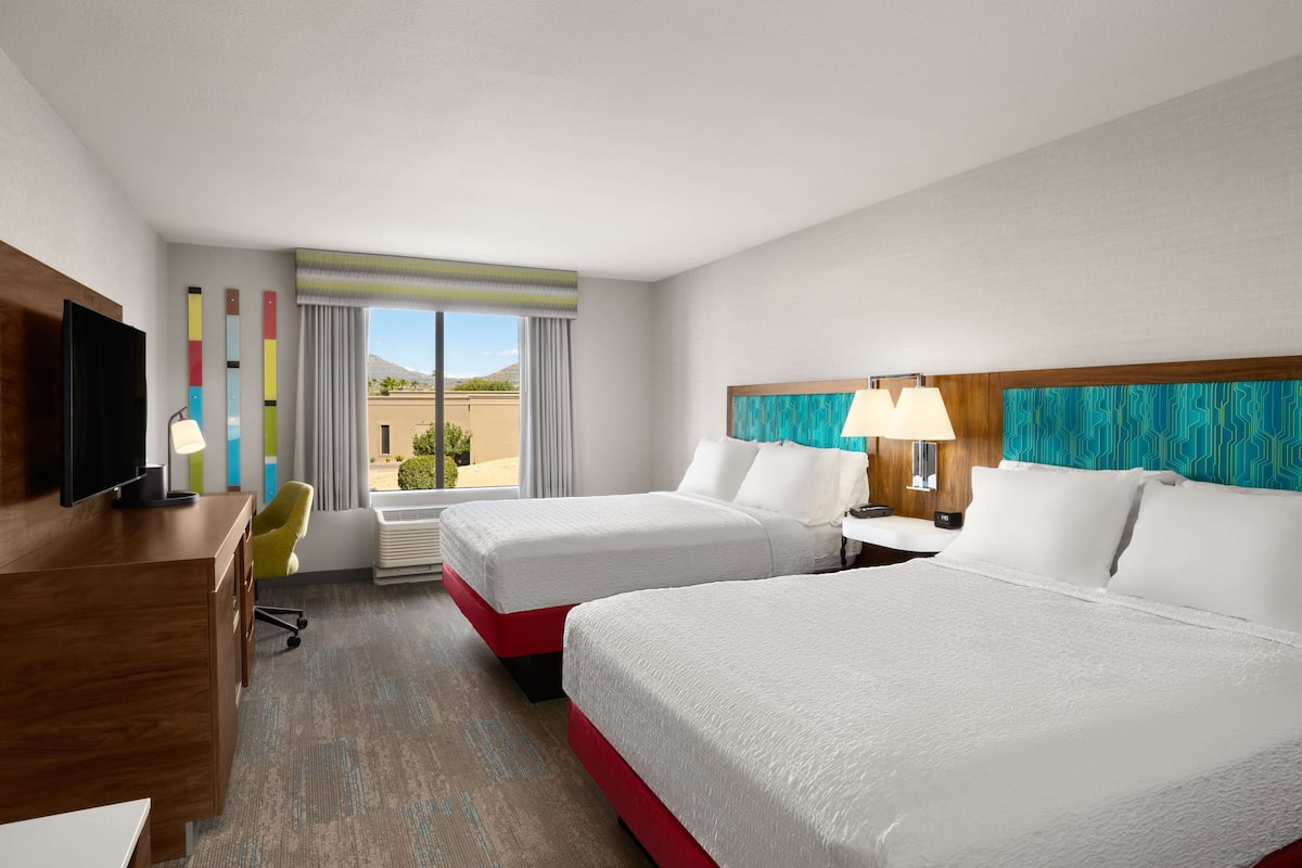 A hotel room with two beds, a desk, a TV, and a view of the outdoors.