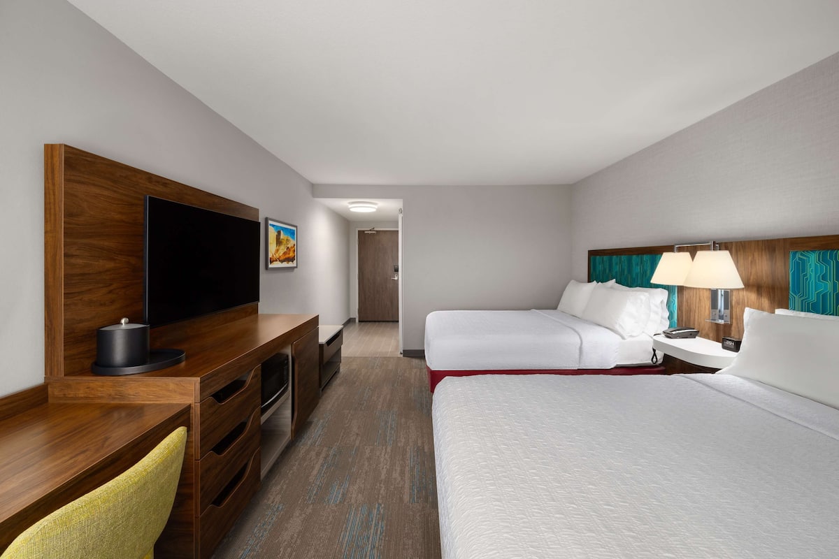 A hotel room with a wooden desk, a flat-screen TV, a bed with white bedding, a nightstand with a lamp, and a door leading to another room.