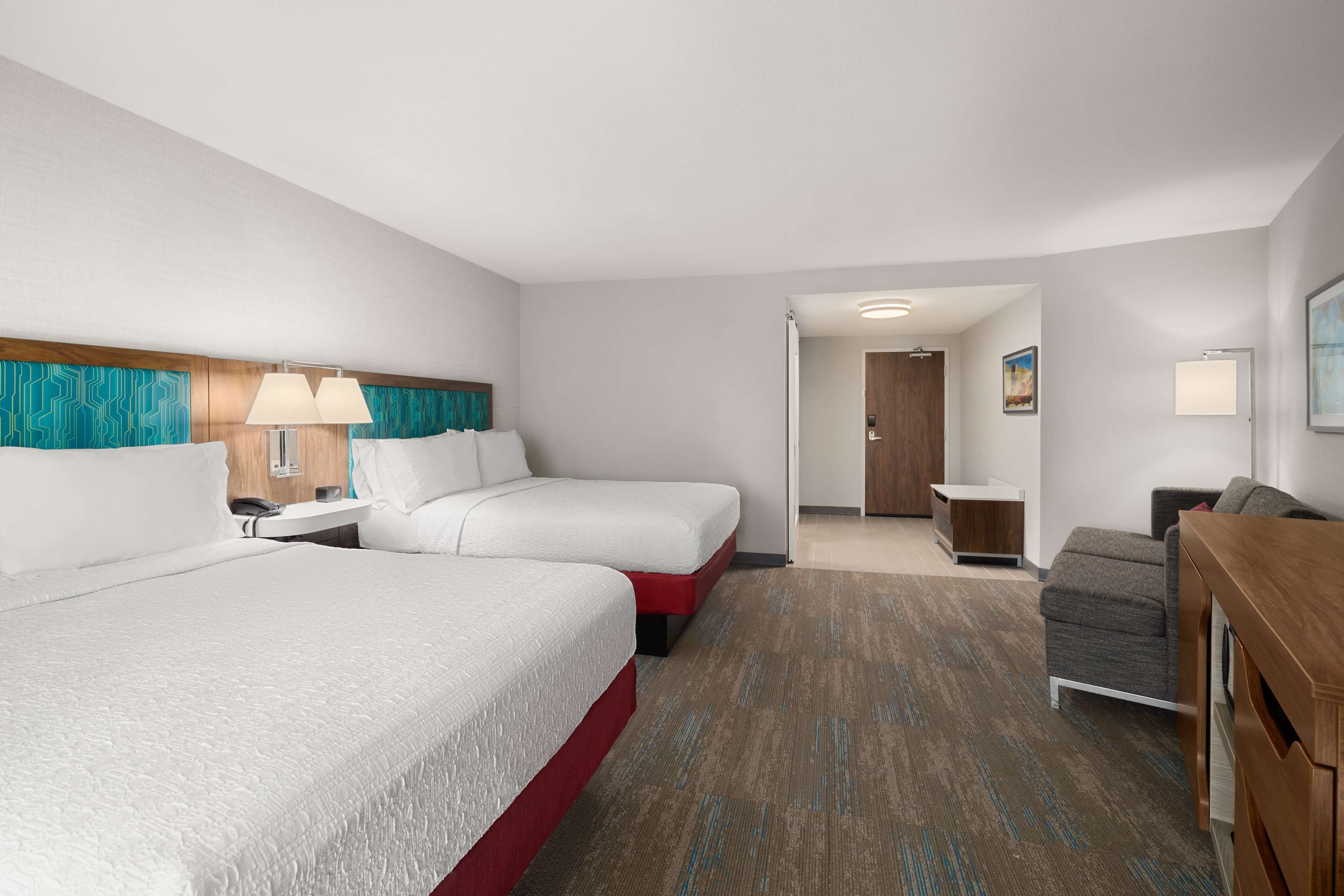 A hotel room with two beds, a desk, a chair, and a small kitchenette.