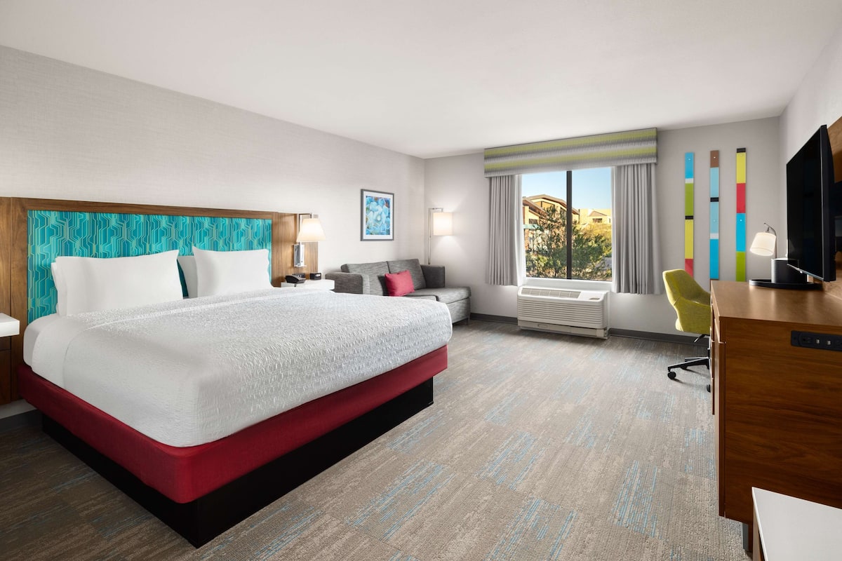 A hotel room with a large bed, a desk, a chair, a TV, and a window with curtains.