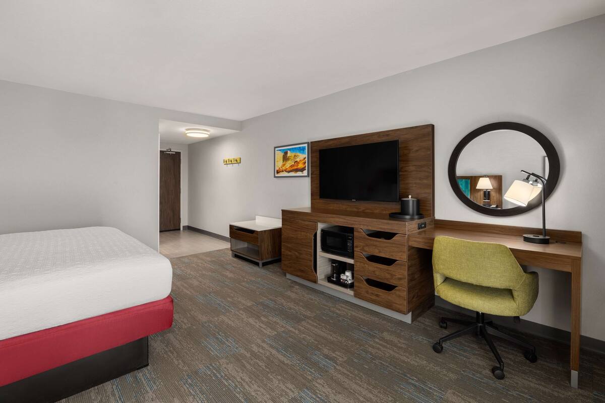 A hotel room with a bed, a desk with a computer, a TV, a mirror, and a small cabinet.