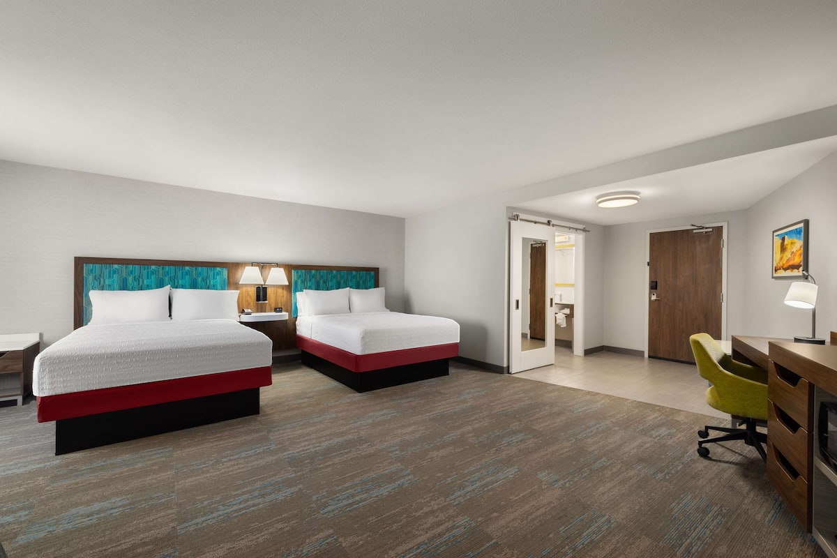 A hotel room with two beds, a desk, a chair, and a door leading to a bathroom.