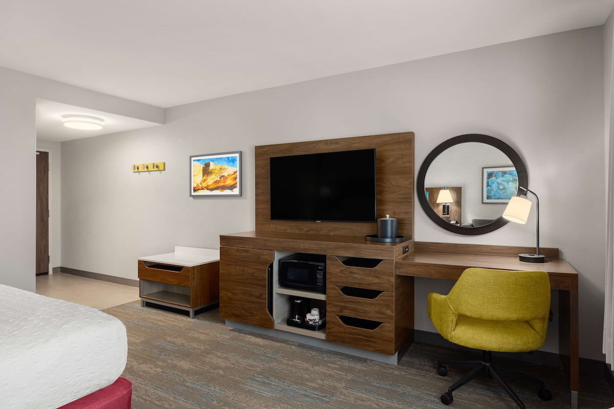 A hotel room with a bed, a desk with a TV, a mirror, and artwork on the walls.
