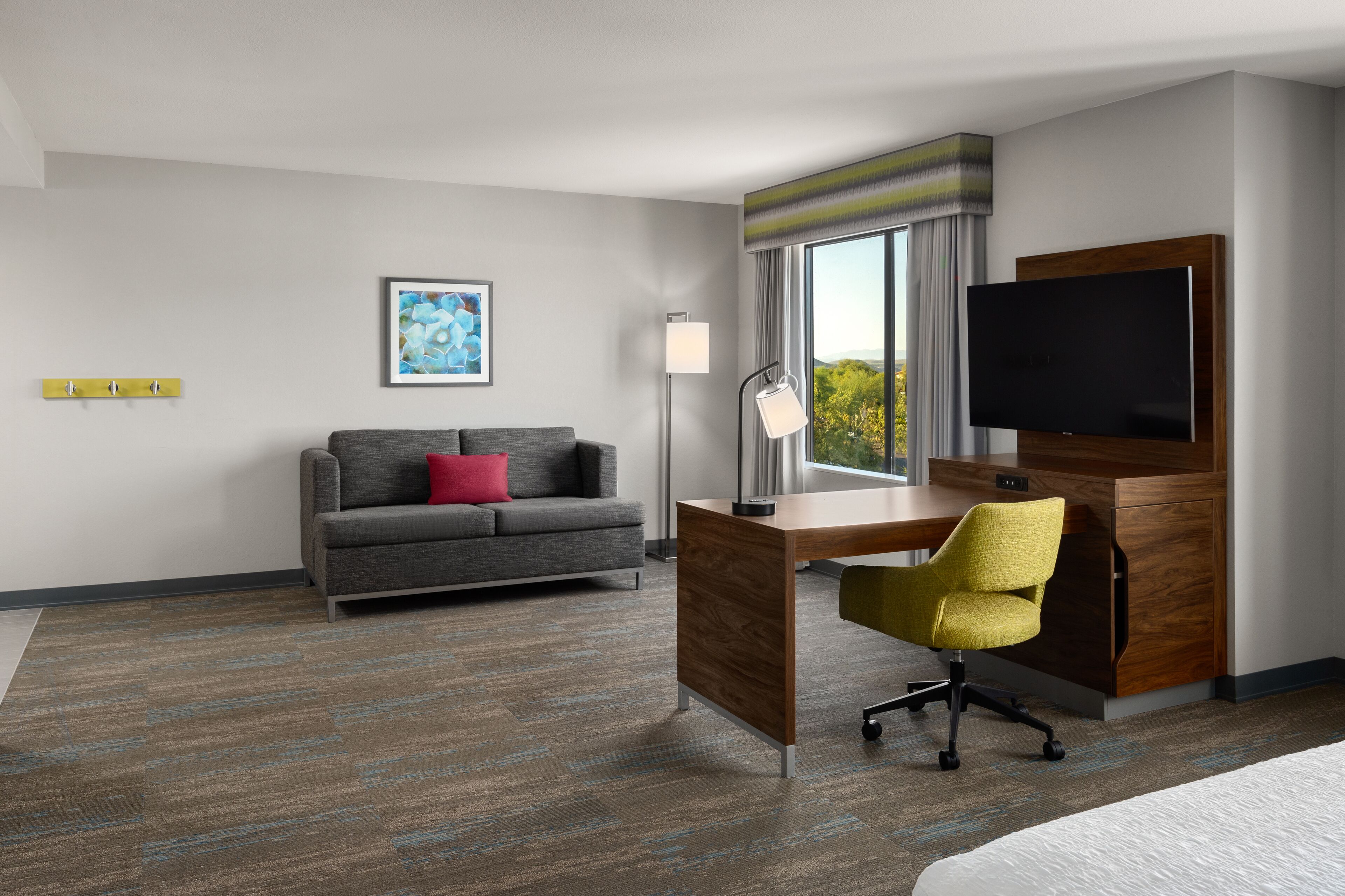 A hotel room with a grey sofa, a yellow chair, a desk, a flat-screen TV, and a large window with a view of trees.