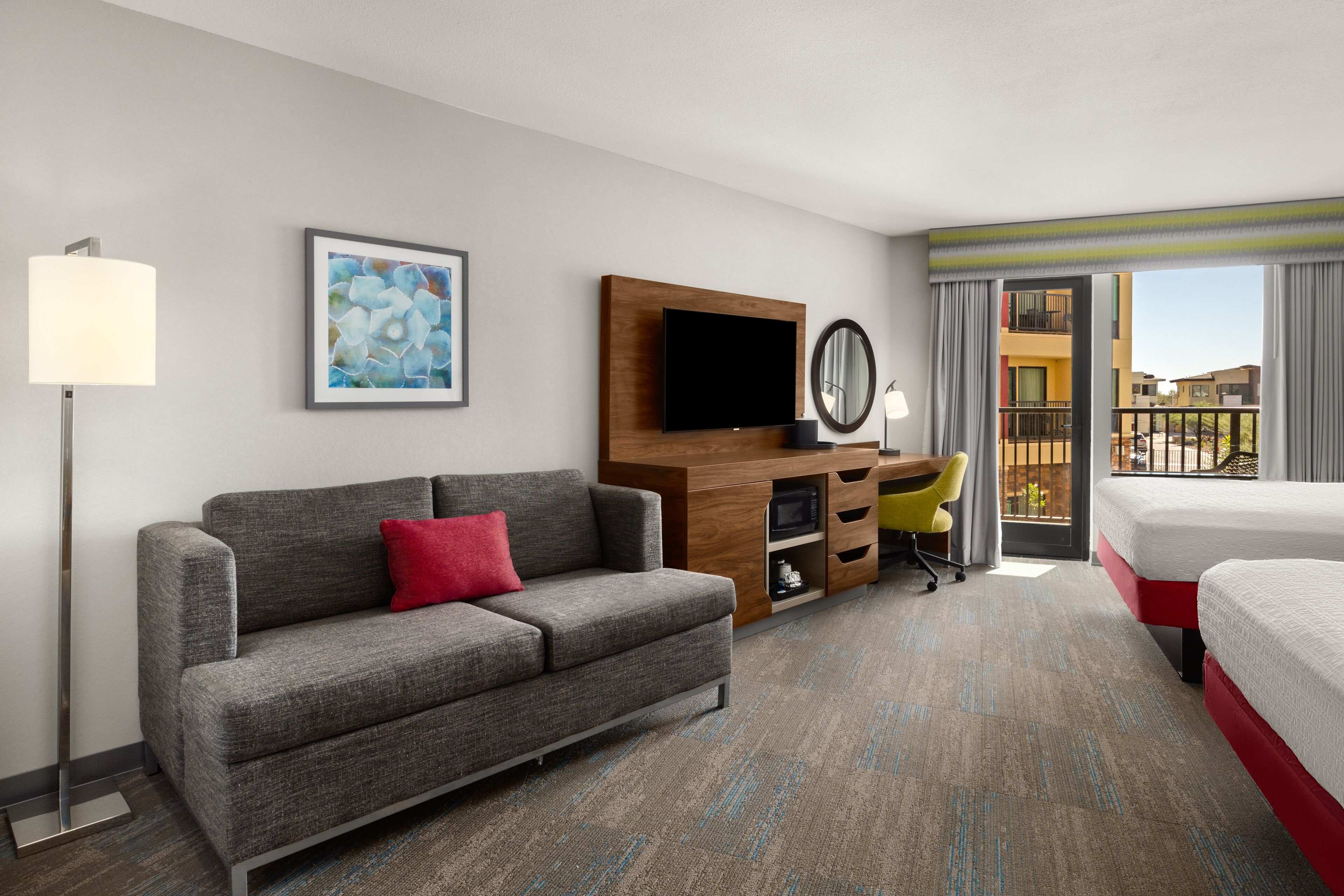 A hotel room with a grey sofa, a bed, a desk with a chair, a flat-screen TV, a mirror, and a balcony with a view of buildings.