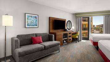 A hotel room with a grey sofa, a bed, a desk with a chair, a flat-screen TV, a mirror, and a balcony with a view of buildings.