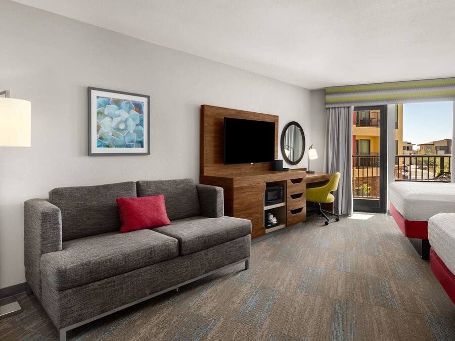 A hotel room with a grey sofa, a bed, a desk with a chair, a flat-screen TV, a mirror, and a balcony with a view of buildings.