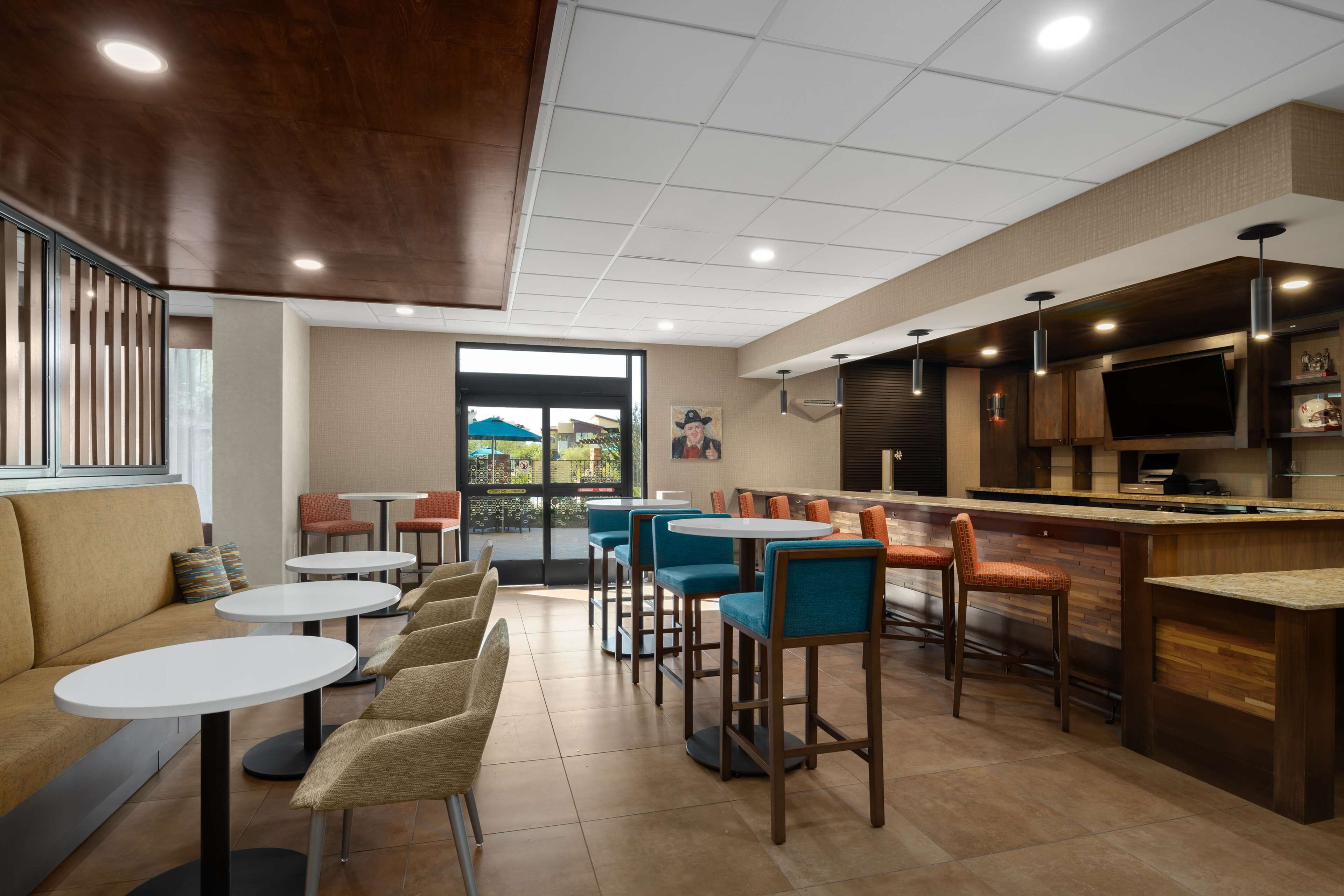 A modern cafe with round tables and bar stools, a counter with a TV, and a view of the outdoors through large windows.