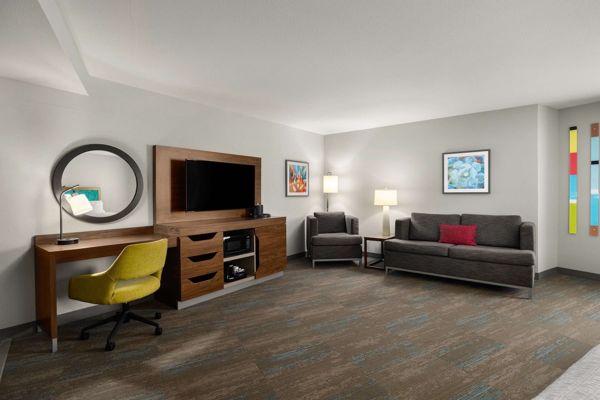 A hotel room with a wooden desk, a yellow office chair, a grey sofa, a TV, and two wall-mounted lamps.