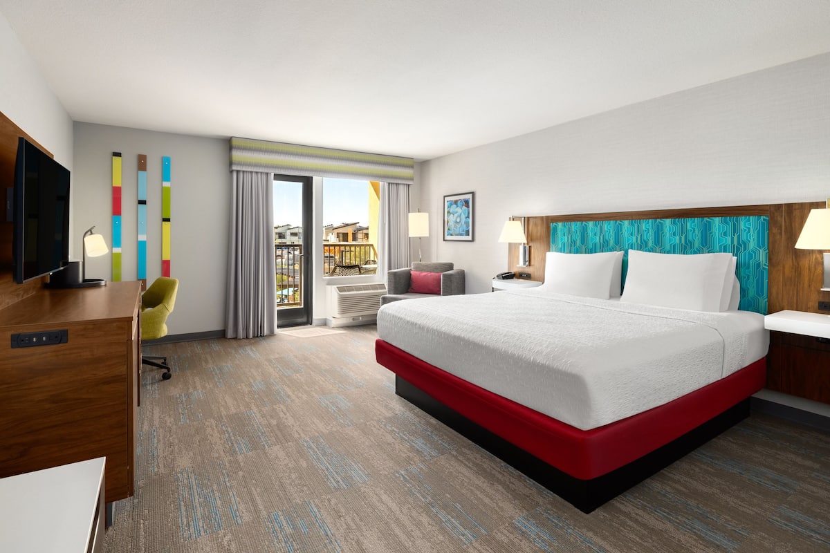 A hotel room with a large bed, a desk, a chair, a TV, and a balcony with a view.