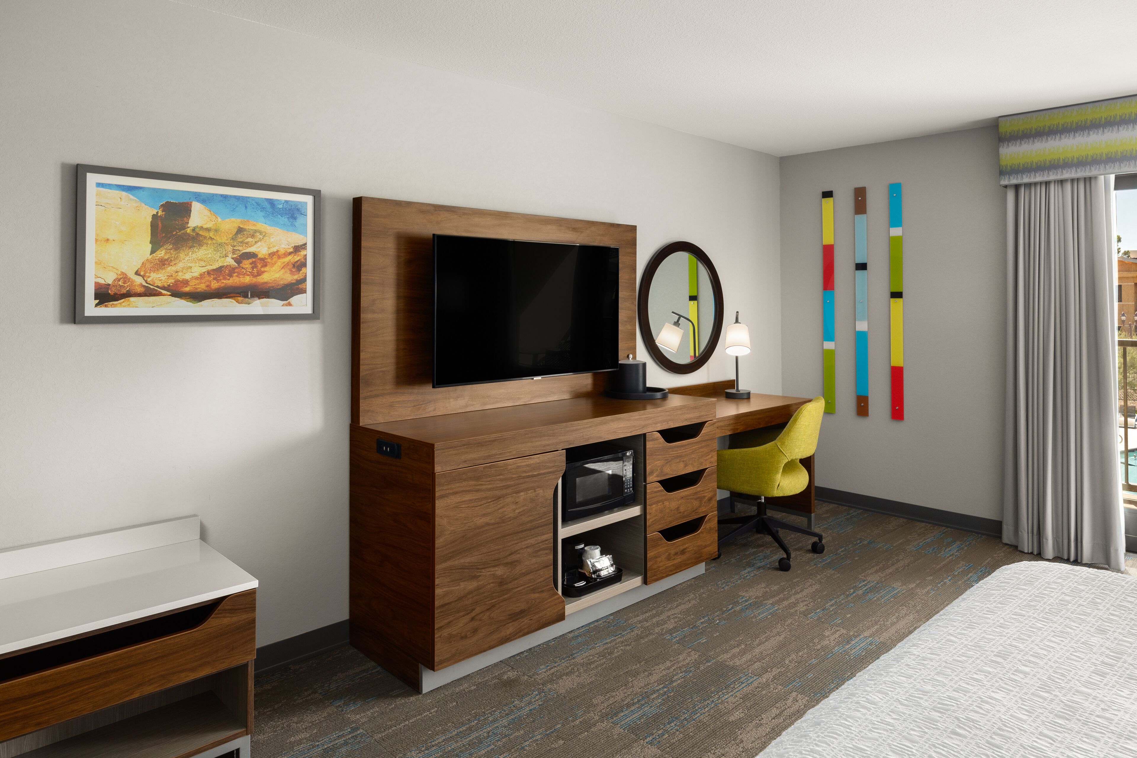 A hotel room with a wooden desk, a flat-screen TV, a mirror, a yellow chair, and a bed.
