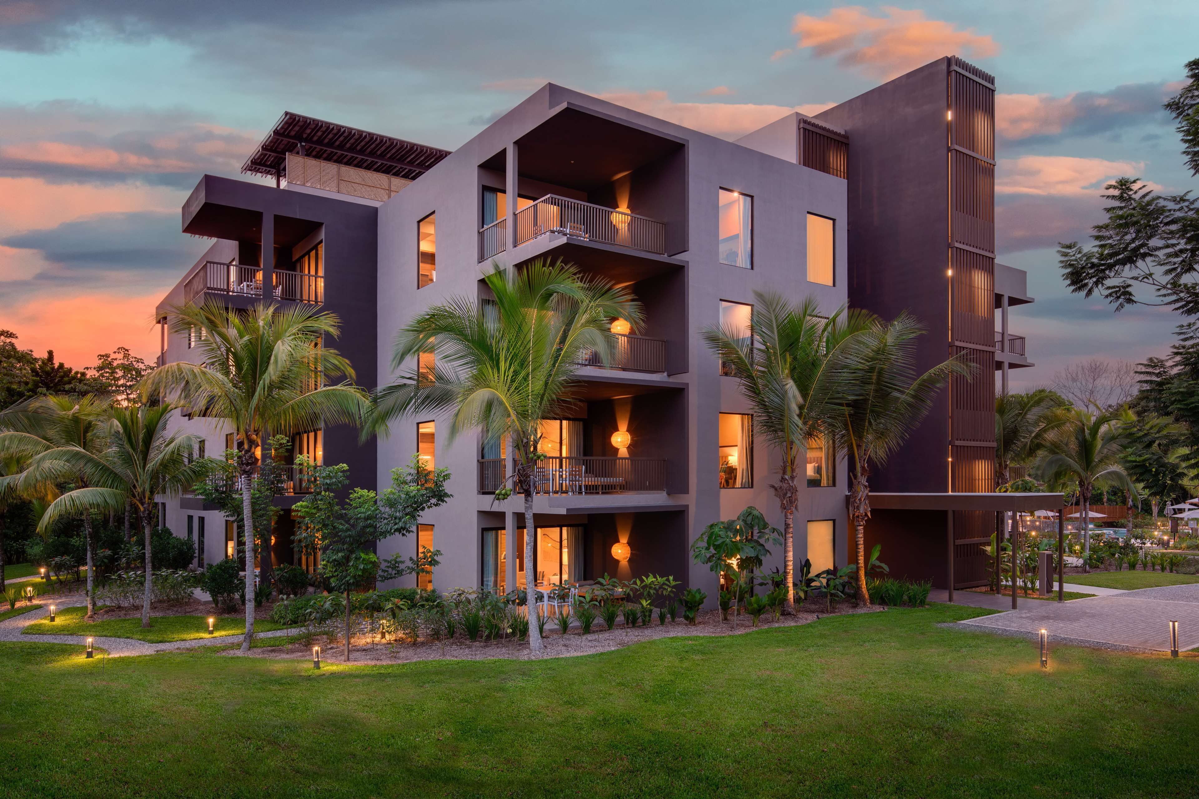 A modern multi-story hotel with balconies, a well-maintained garden, and a sunset sky.