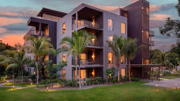 A modern multi-story hotel with balconies, a well-maintained garden, and a sunset sky.