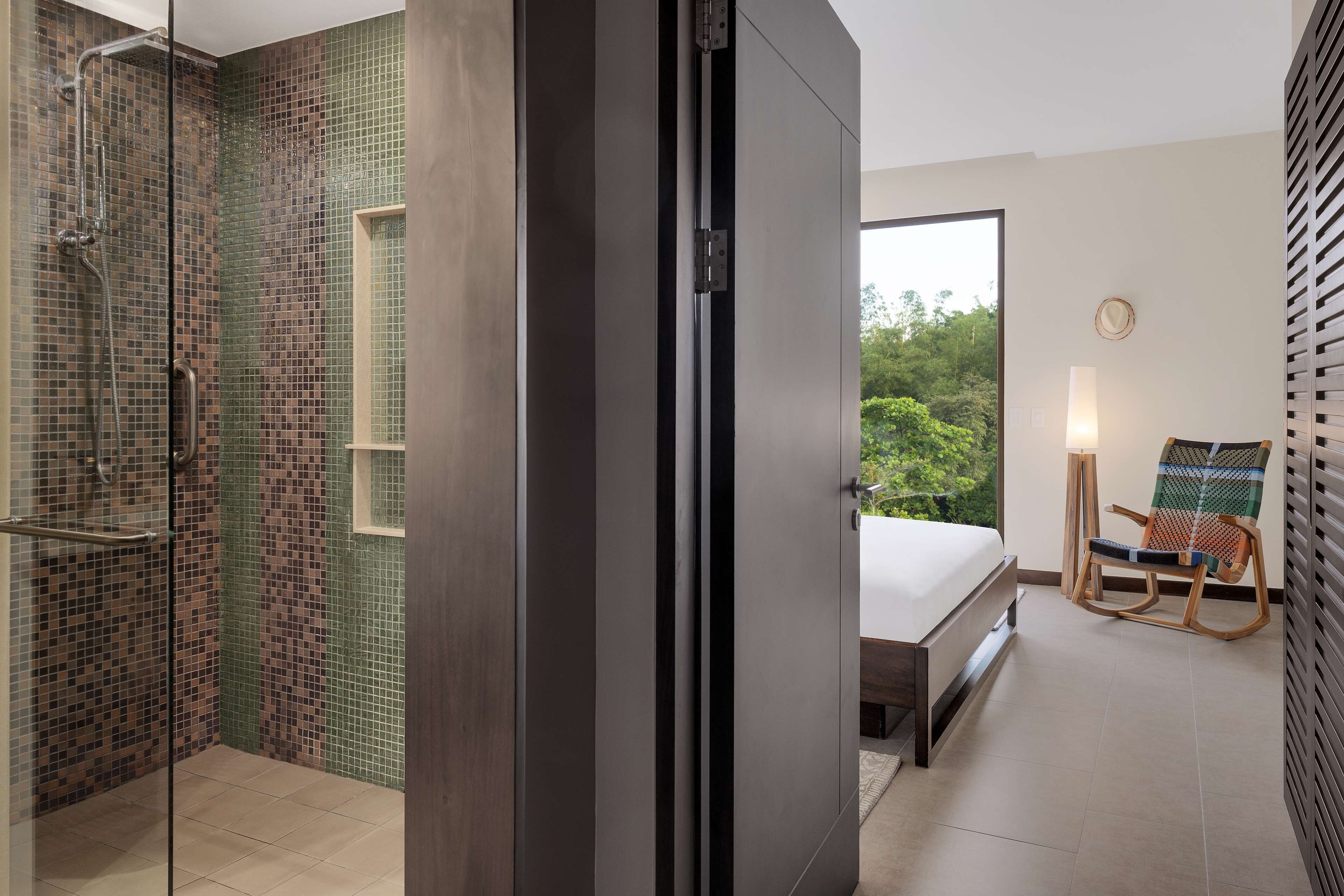 Uma casa de banho moderna com chuveiro, porta de vidro e vista para um quarto com cama, cadeira e janela.