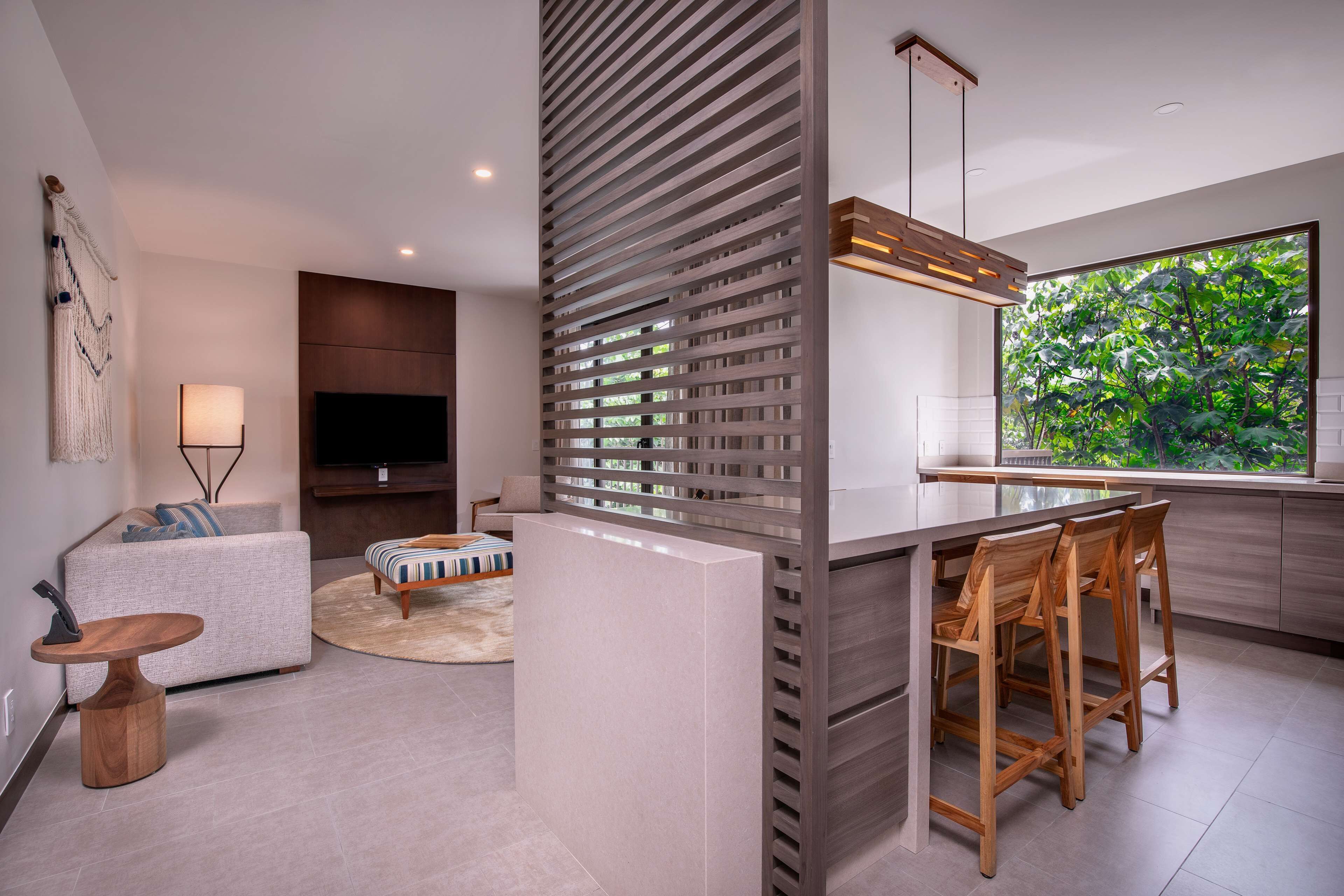 A modern living room with a sofa, a round coffee table, and a TV. There's a wooden partition with a window showing greenery outside.