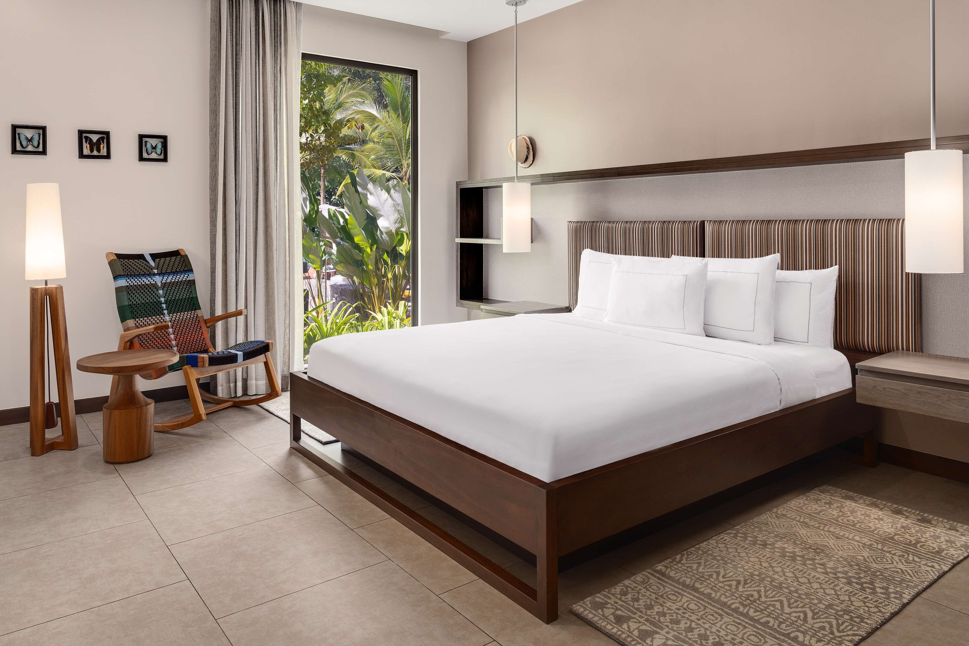 A hotel room with a large bed, a wooden nightstand, a small table with a lamp, a rocking chair, and a view of greenery through the window.