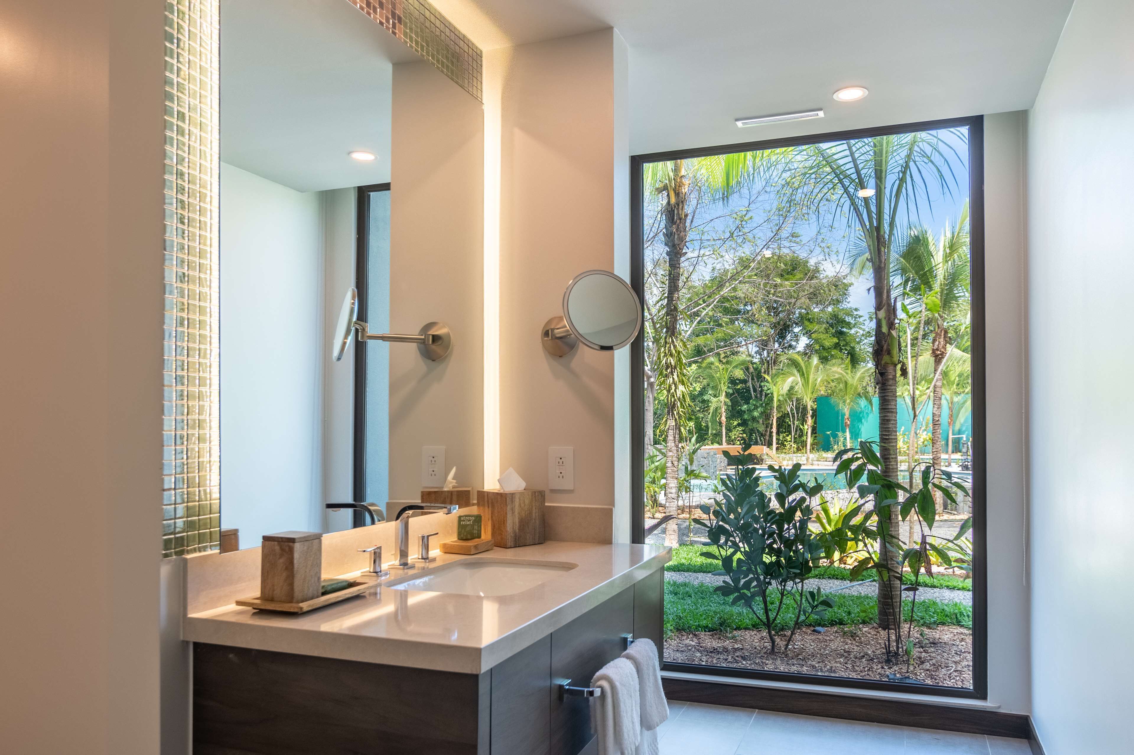 A modern bathroom with a large mirror, a sink, and a view of a tropical garden through a large window.
