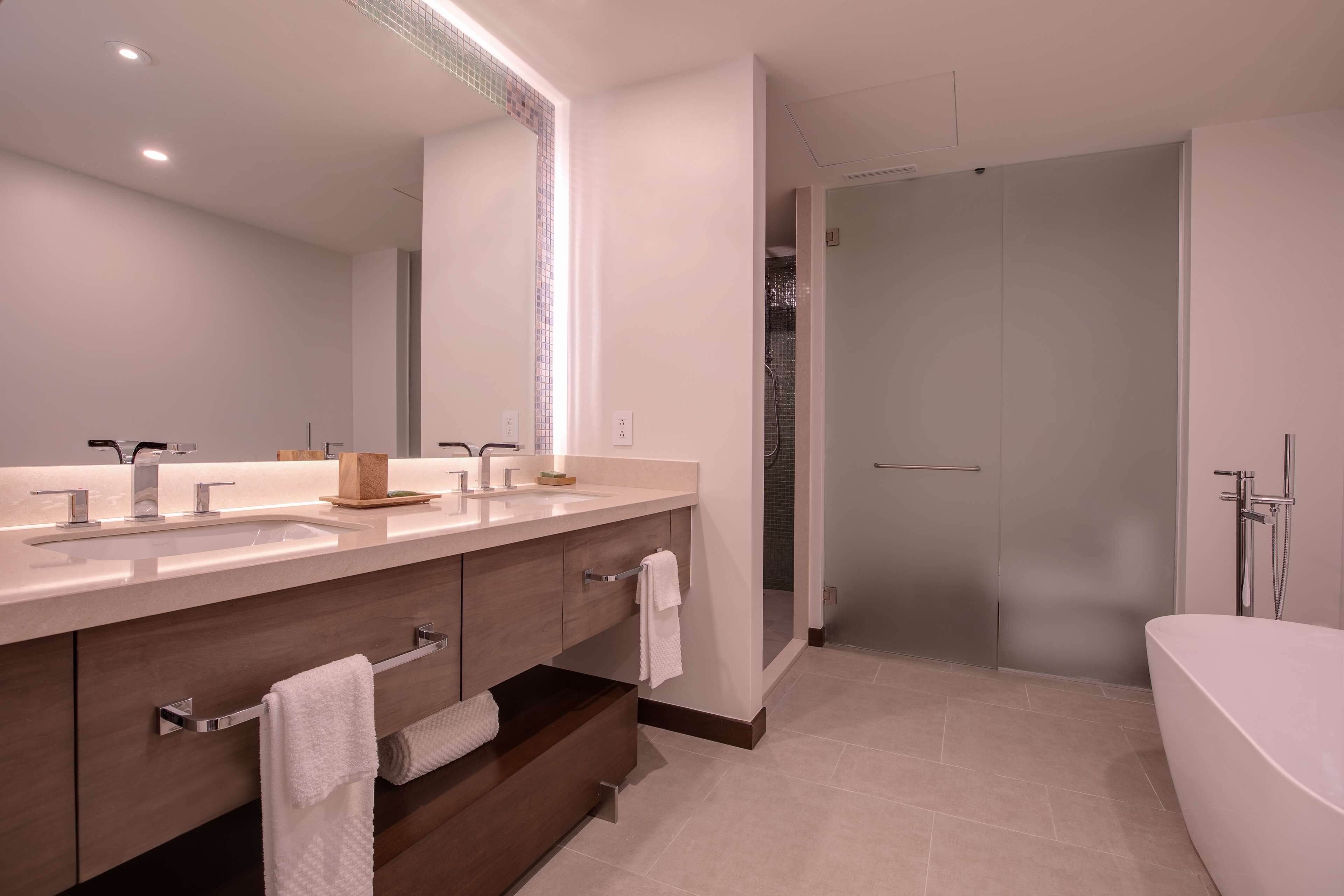 A modern bathroom with a large mirror, two sinks, a vanity with drawers, a bathtub, and a glass-enclosed shower area.