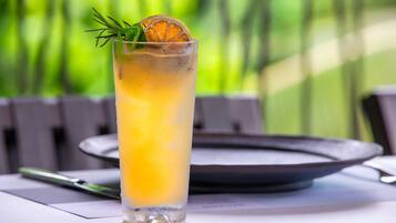 Un vaso alto con una bebida amarilla, adornado con una ramita de romero y una rodaja de naranja, sobre una mesa con un plato y un bolígrafo.