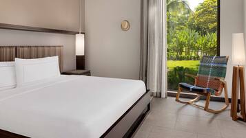 A hotel room with a large bed, a wooden rocking chair, a nightstand, a lamp, and a view of greenery through a window.