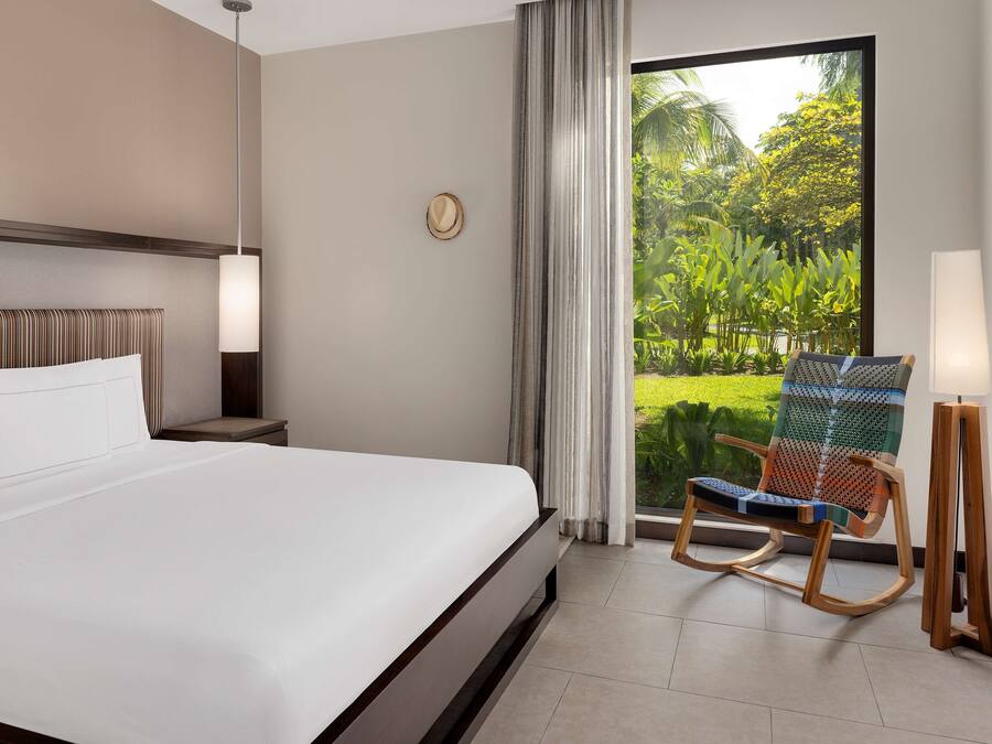 A hotel room with a large bed, a wooden rocking chair, a nightstand, a lamp, and a view of greenery through a window.