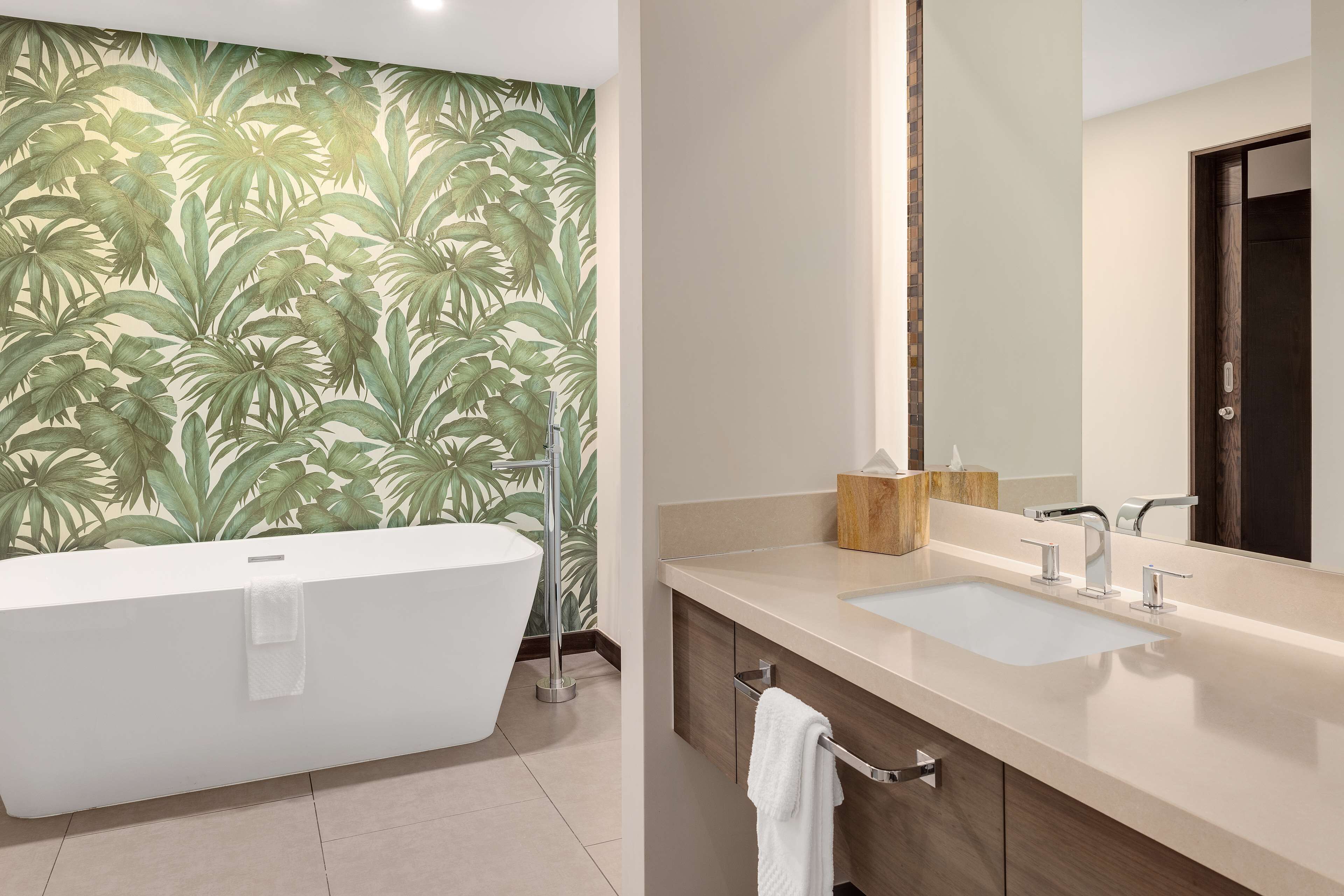 A bathroom with a large bathtub, a vanity with a mirror, and a wall with a tropical leaf pattern.