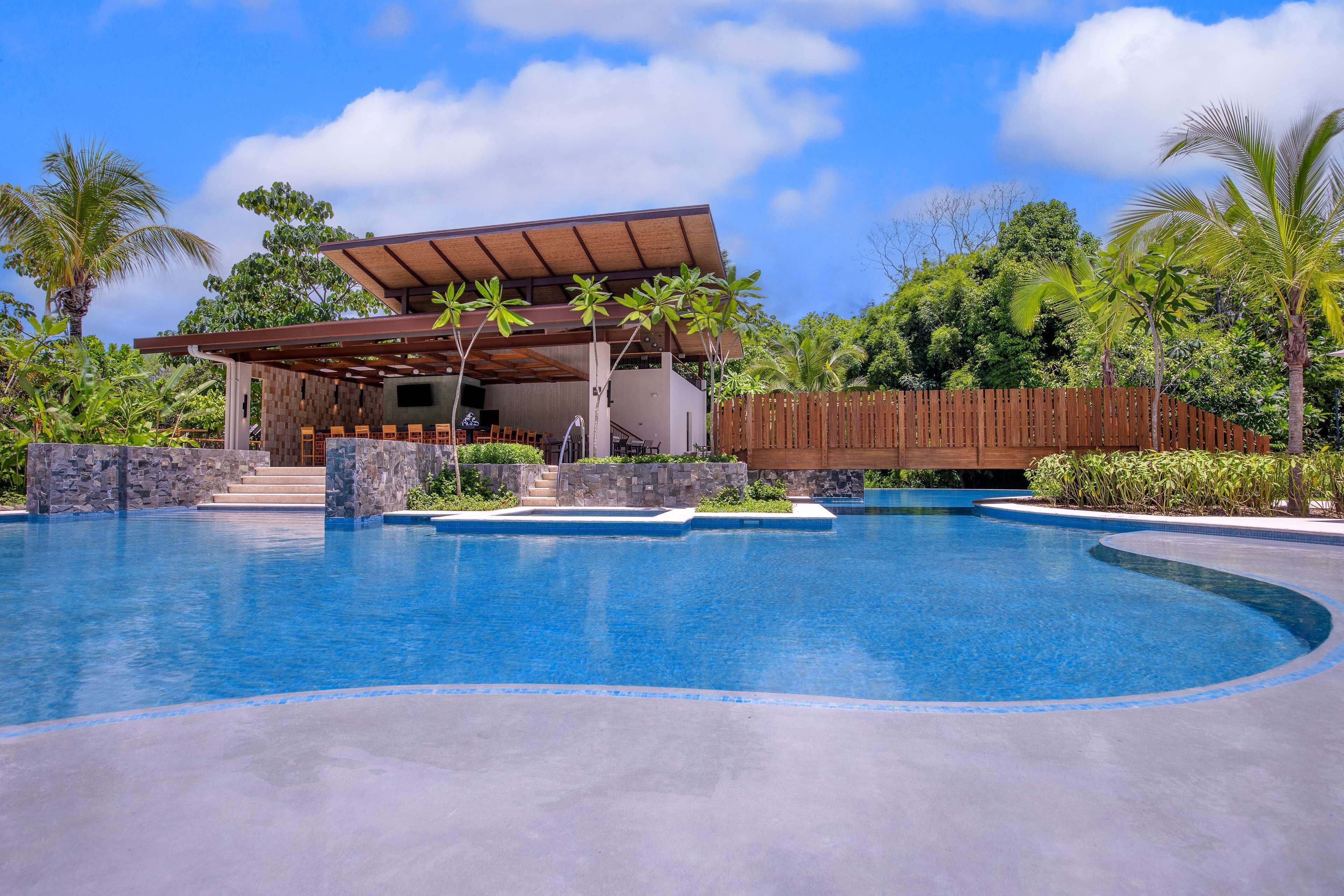 Un área de piscina moderna con una zona de estar cubierta, escalones de piedra y exuberante vegetación tropical.