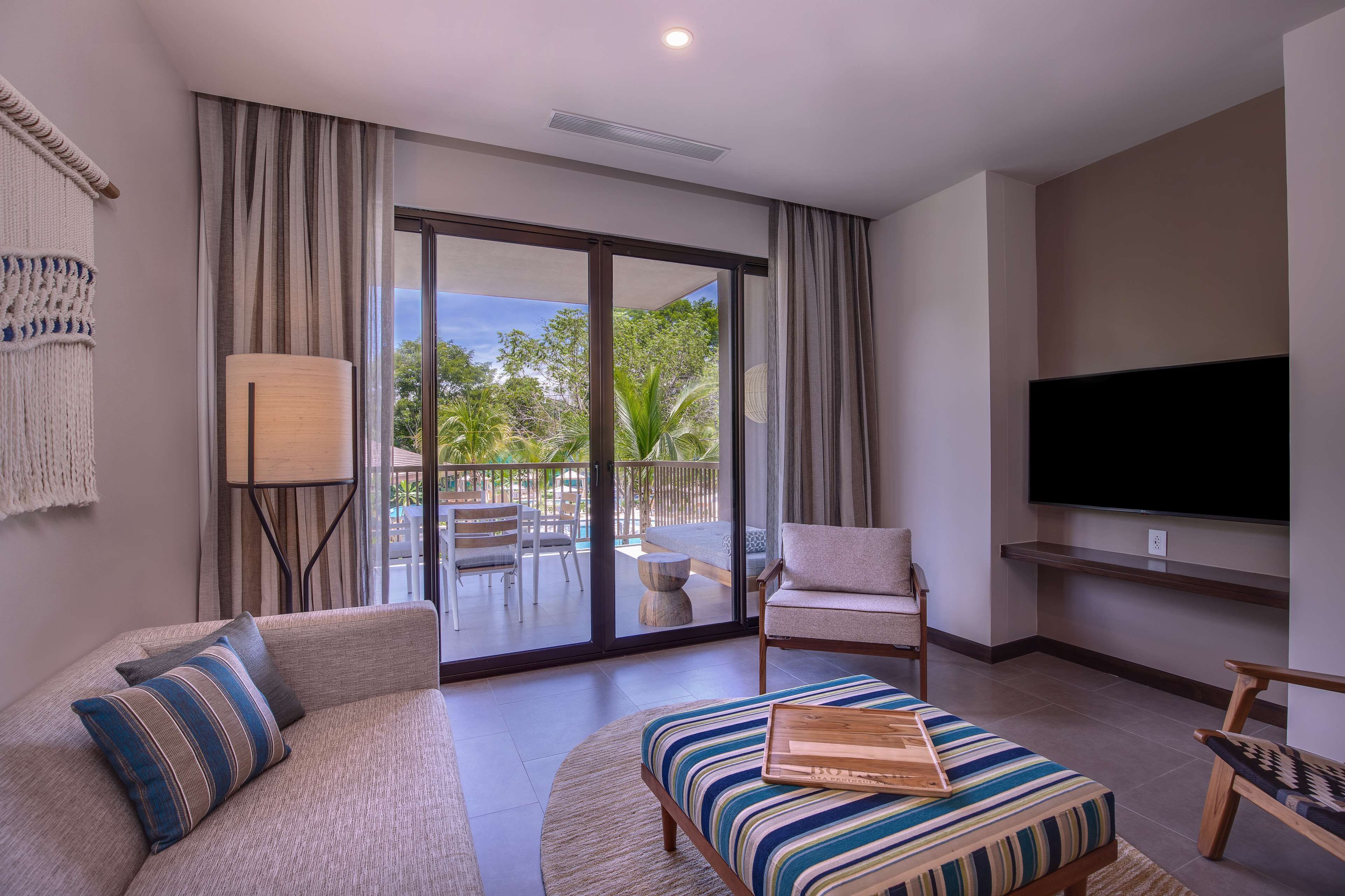 A modern living room with a sofa, ottoman, and armchair, a flat-screen TV, and a balcony with outdoor furniture.