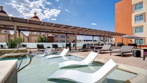 Ein Poolbereich auf dem Dach mit Liegestühlen und Sonnenschirmen, umgeben von einer überdachten Terrasse und einem Gebäude im modernen Stil.