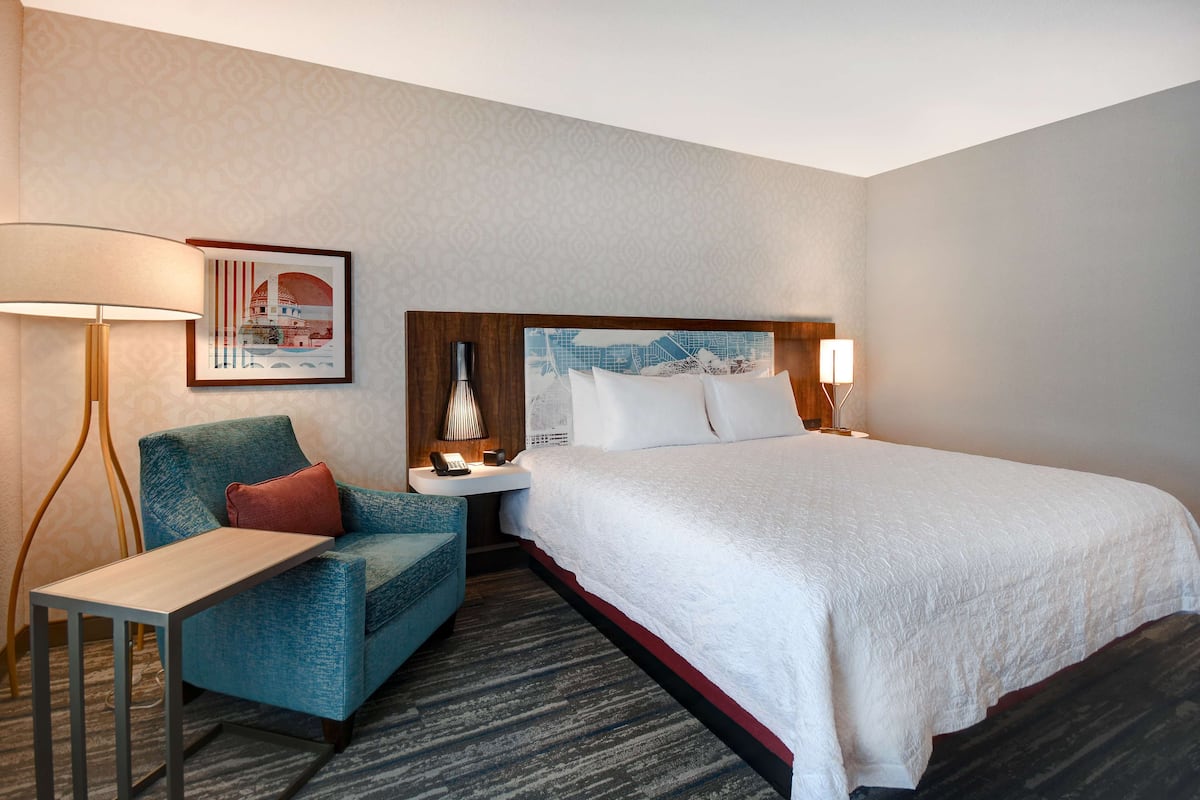 A hotel room with a large bed, a desk, a chair, and a nightstand with a lamp.