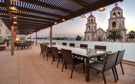 An outdoor patio with a long table and wicker chairs, overlooking a historic church with two bell towers.