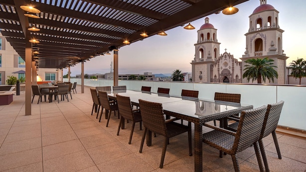 Eine Terrasse im Freien mit einem langen Tisch und Korbstühlen, mit Blick auf eine historische Kirche mit zwei Kirchtürmen.