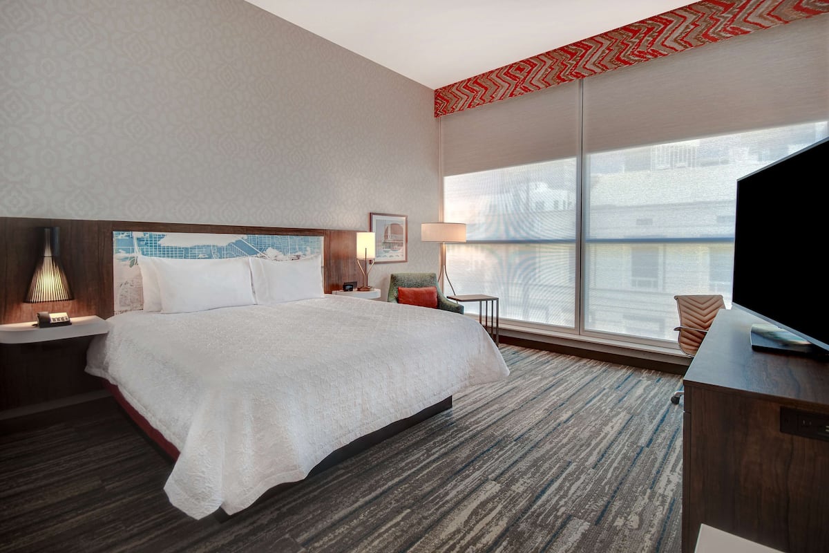 A hotel room with a large bed, a desk, a chair, a TV, a window with a view, and a patterned ceiling.