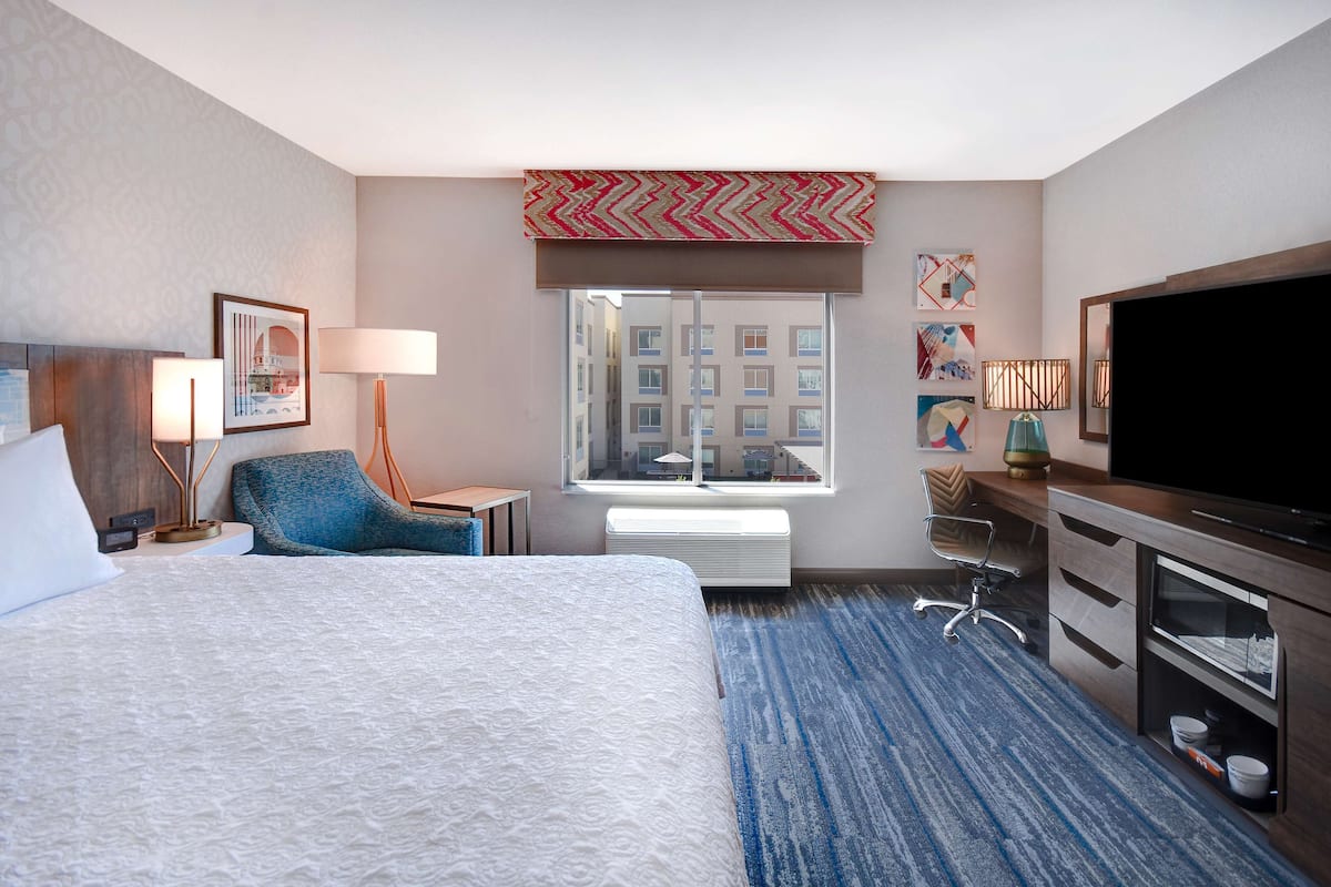 A hotel room with a large bed, a desk, a television, and a window with a view of buildings.