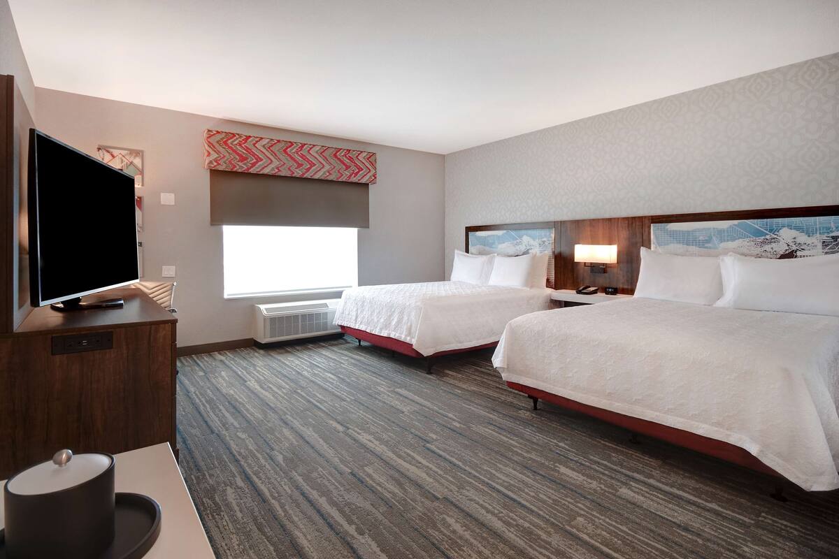 A hotel room with two beds, a flat-screen TV, a desk with a lamp, and a large window.