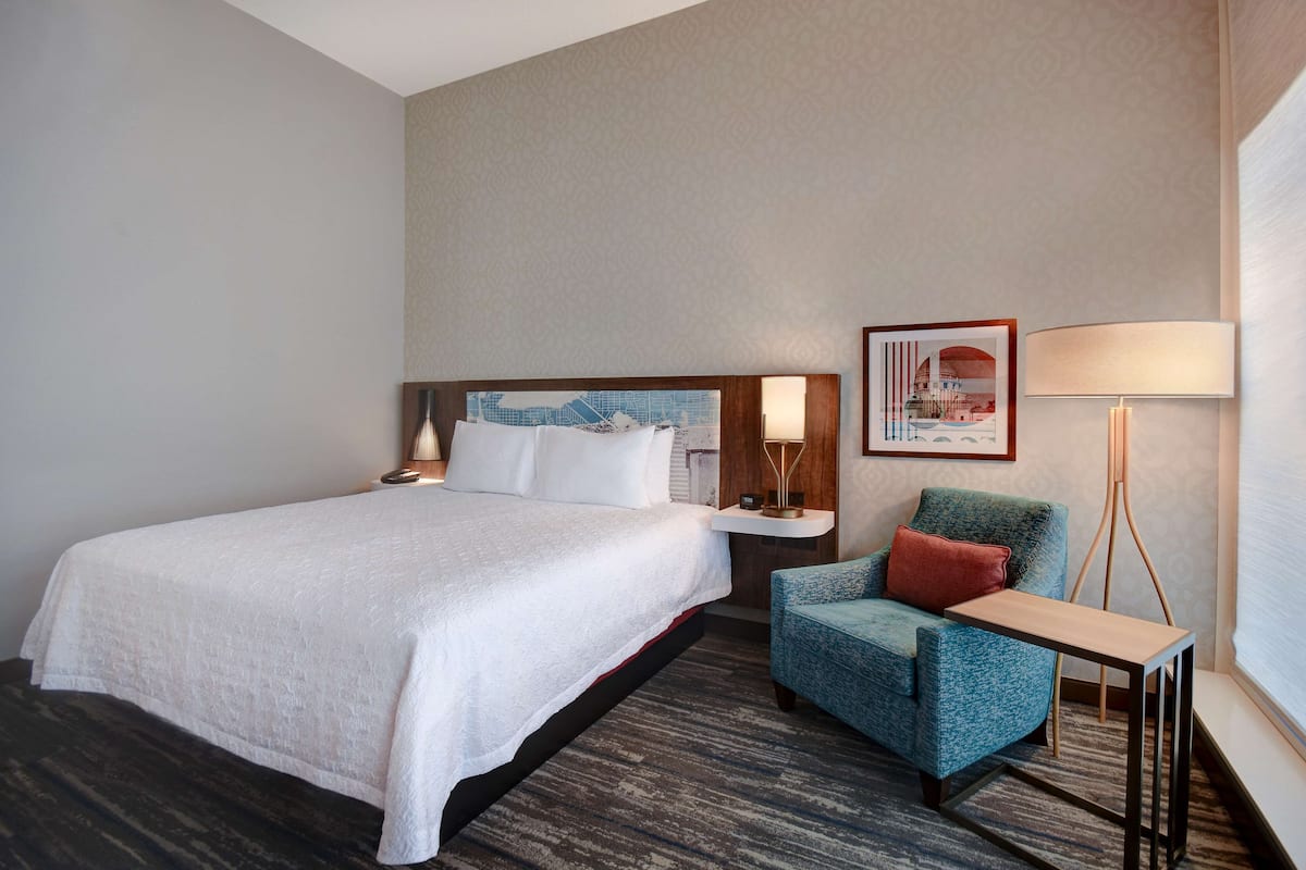 A hotel room with a large bed, a small desk, a chair, a lamp, and a framed picture on the wall.