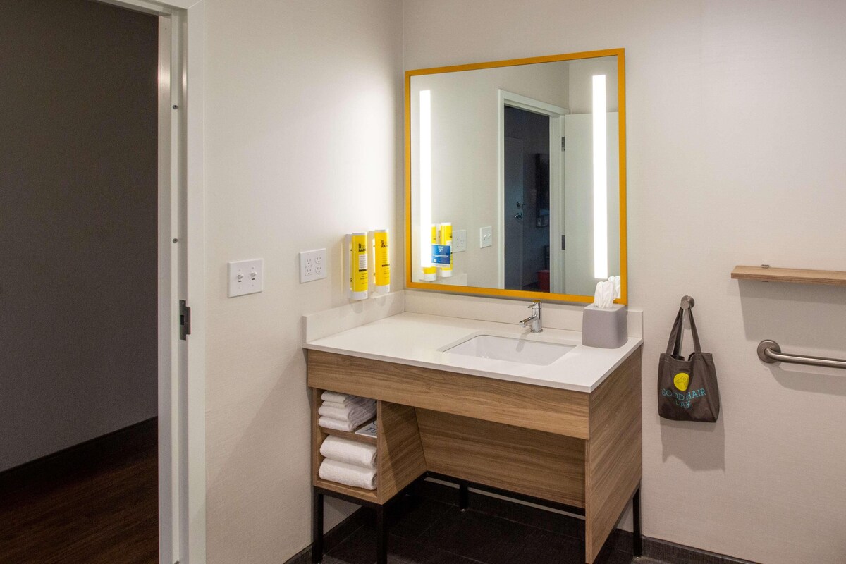Une salle de bain avec un lavabo blanc, un meuble-lavabo en bois, un miroir avec un cadre jaune, un porte-serviettes et une petite étagère contenant des articles de toilette.